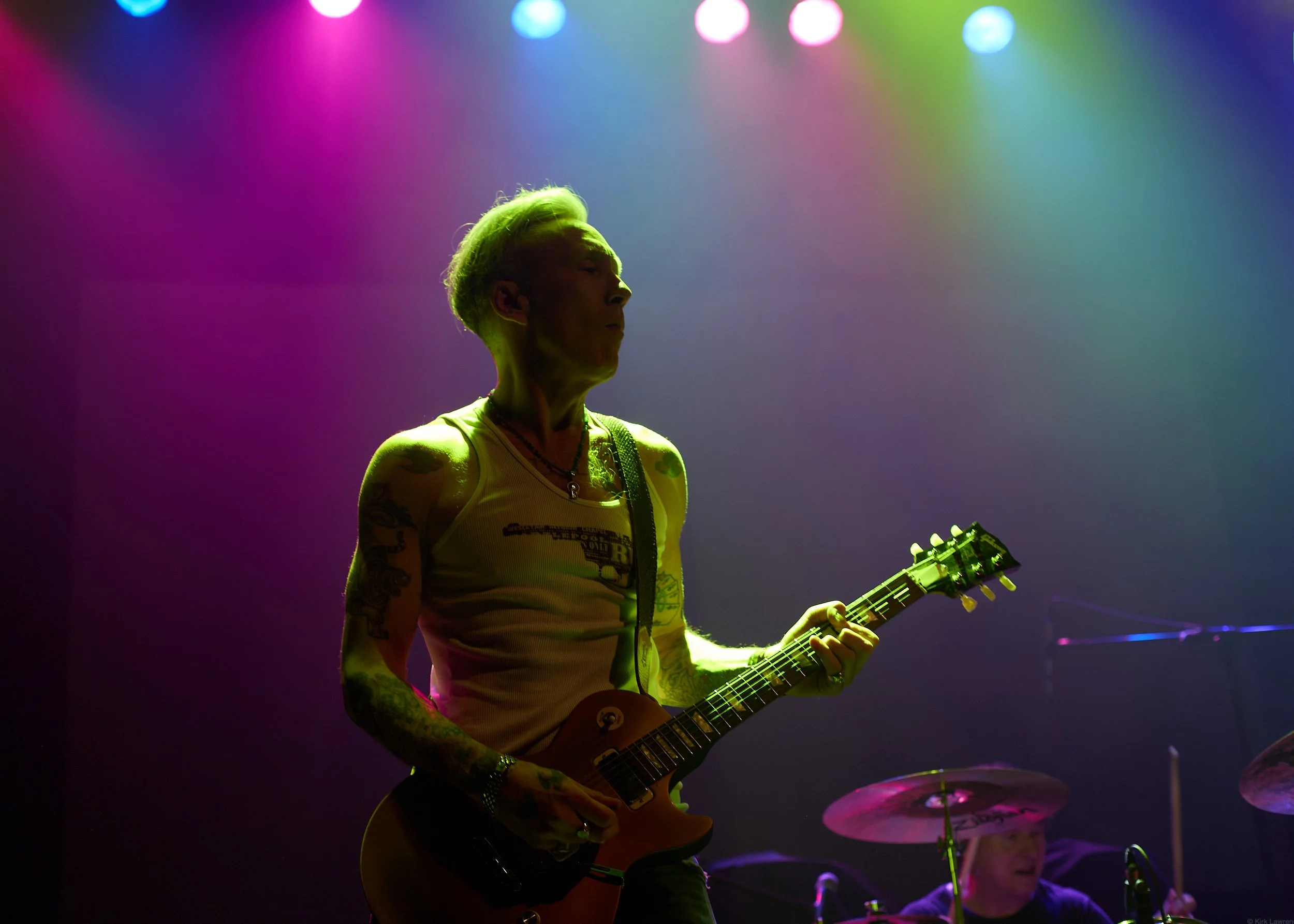 A person with green hair playing an electric guitar on stage under colorful lights, wearing a sleeveless shirt with tattoos on their arms.