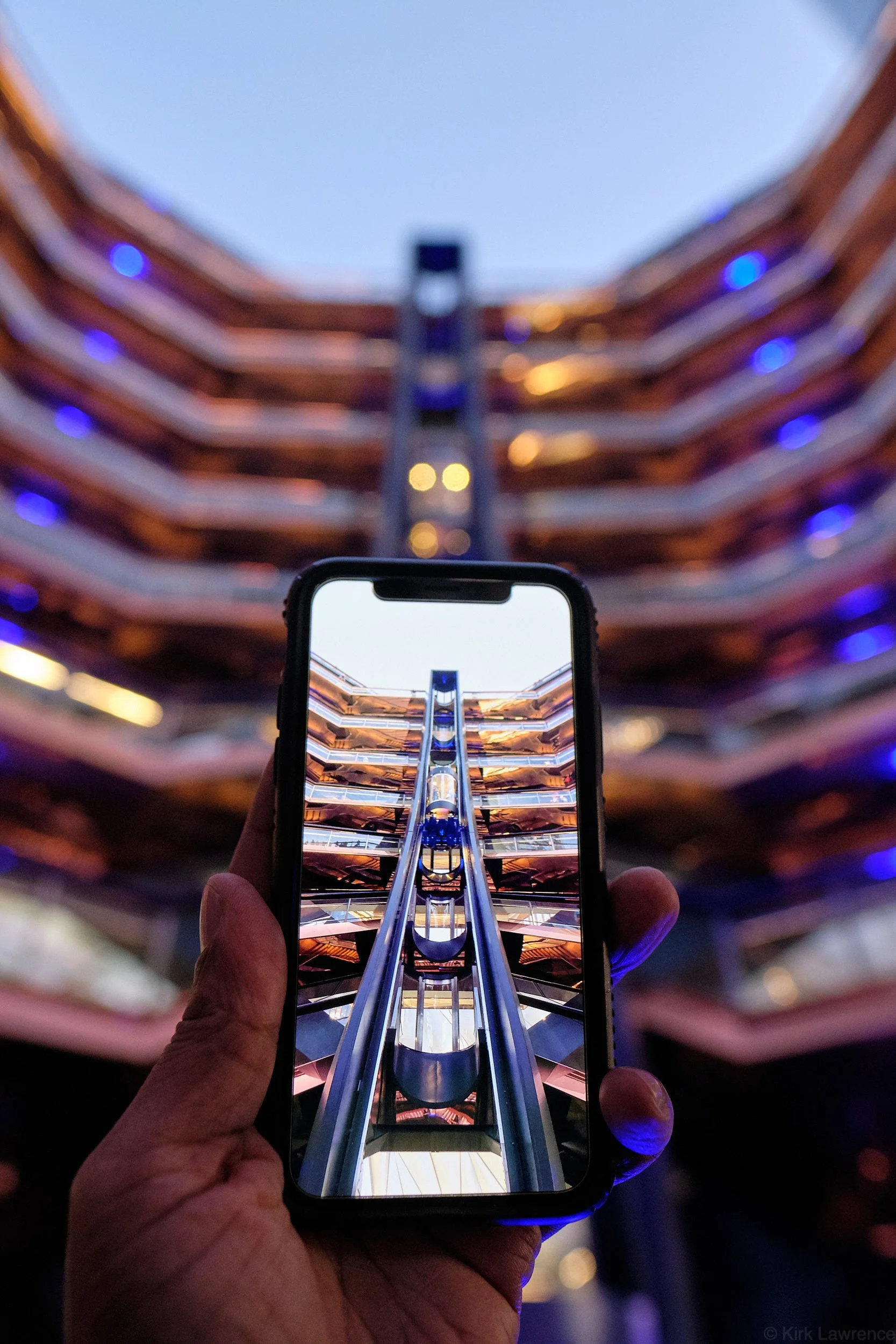A person holding a smartphone captures a photo of the Vessel in New York City with multiple curved balconies with blue lights. The phone screen displays the same scene in focus, creating a visual effect of a frame within a frame.