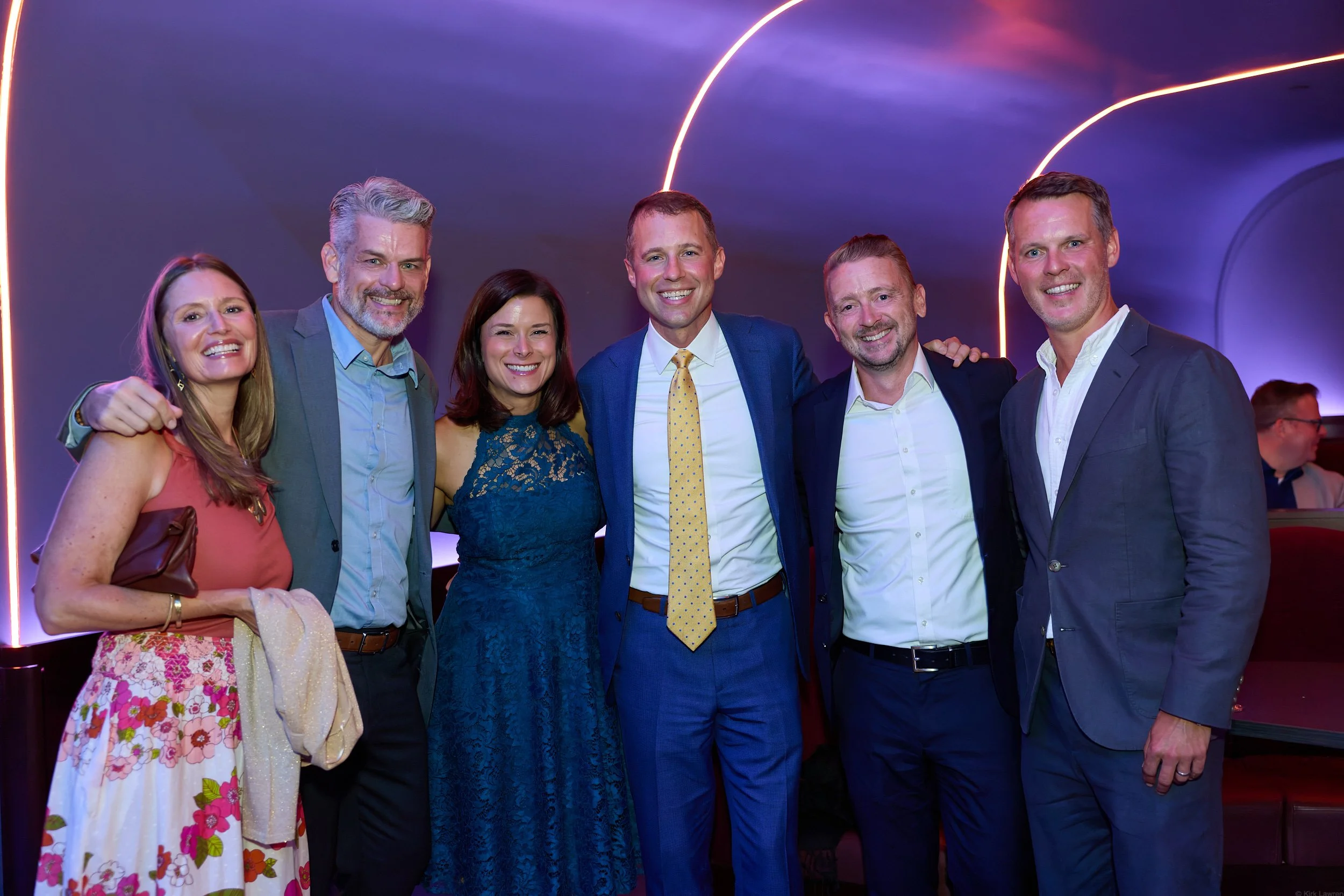 Group of six people smiling at a formal event, standing close together in front of a purple background with neon lighting.