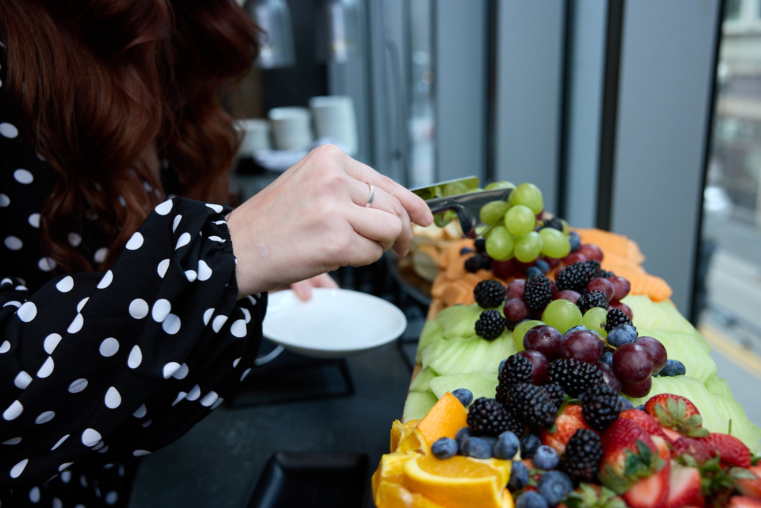 Person with red hair in a black shirt with white polka dots using tongs to pick grapes from a fruit display, with strawberries, blackberries, and sliced oranges on a table.