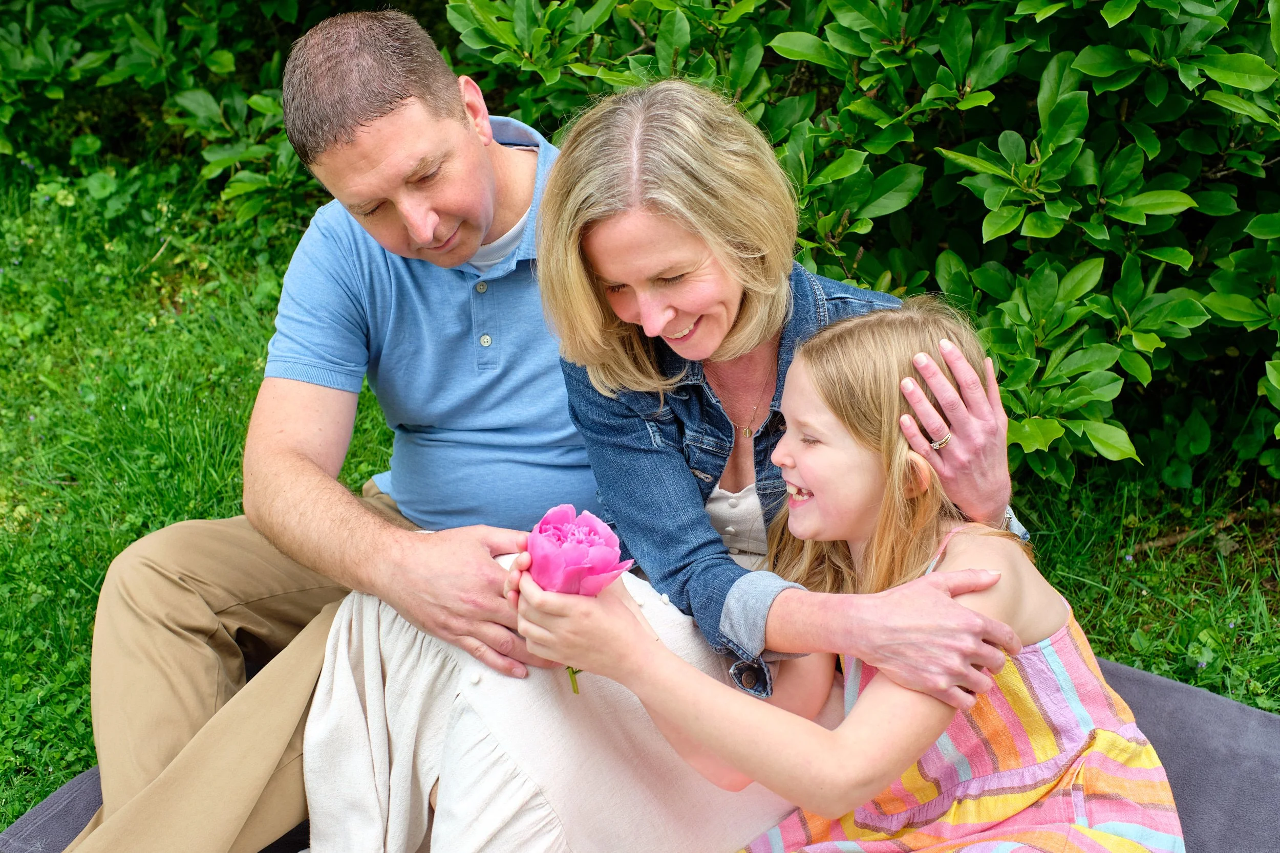 A family of four, including a man, woman, and two young girls, sitting outdoors in a garden with green bushes, sharing a joyful moment as they look at a pink flower.