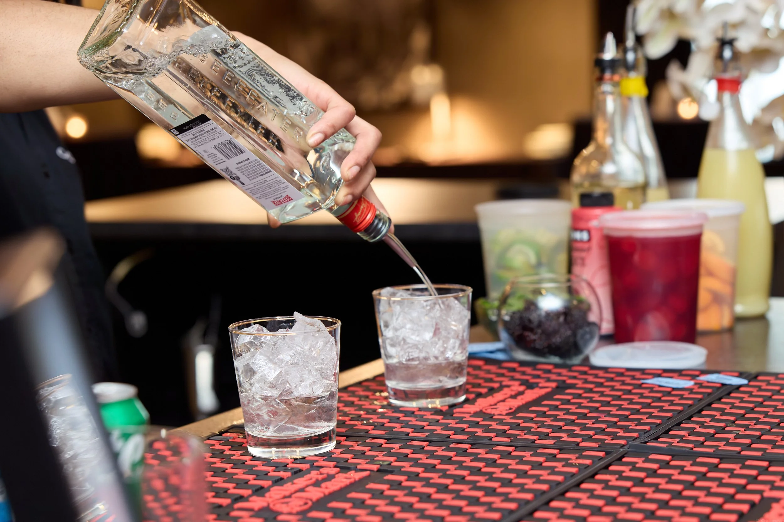 A person pouring clear liquor over ice into a glass at a bar or restaurant, with various bottles and containers in the background.
