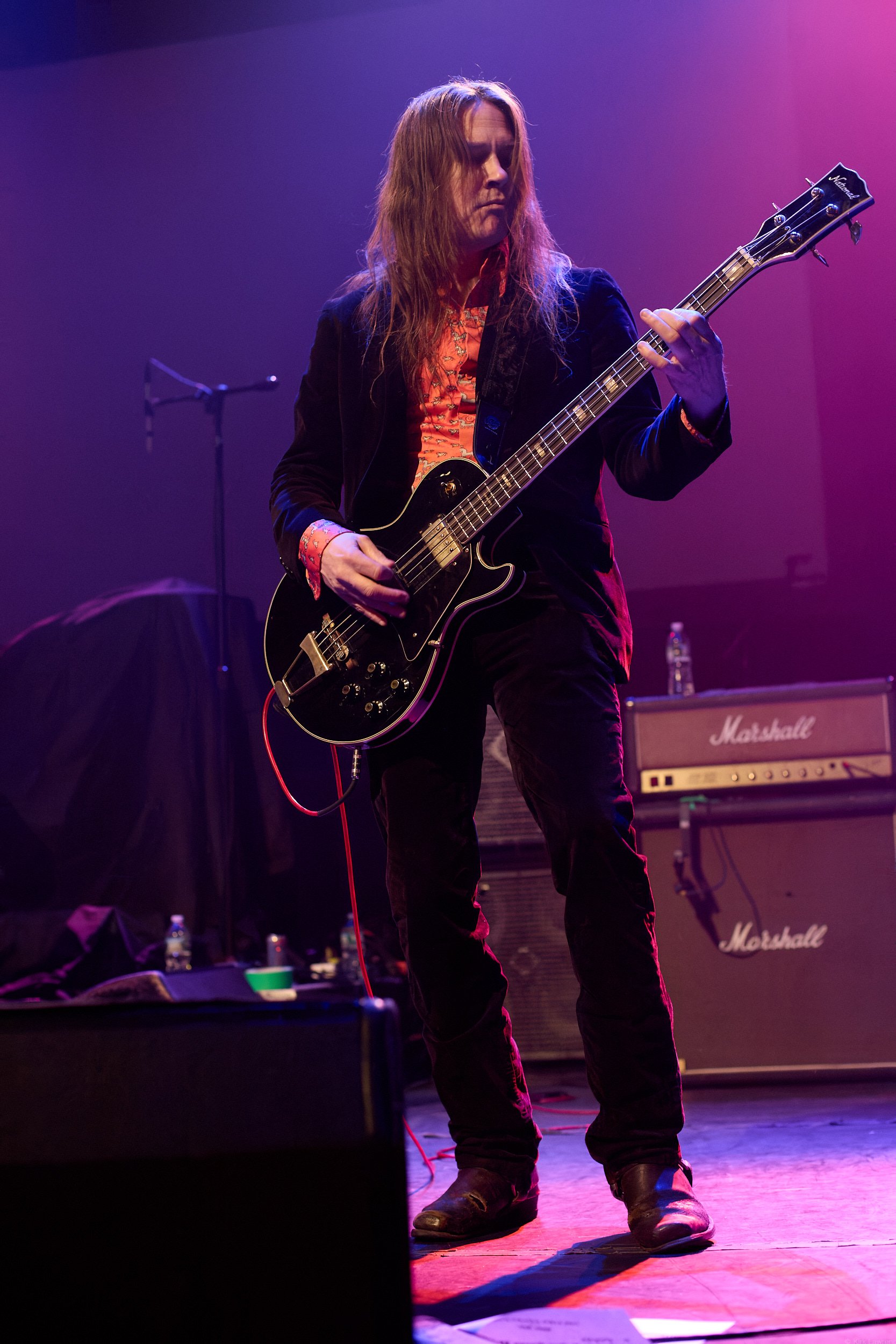 A musician with long hair playing an electric guitar on stage under pink and purple lights, with Marshall amplifiers in the background.