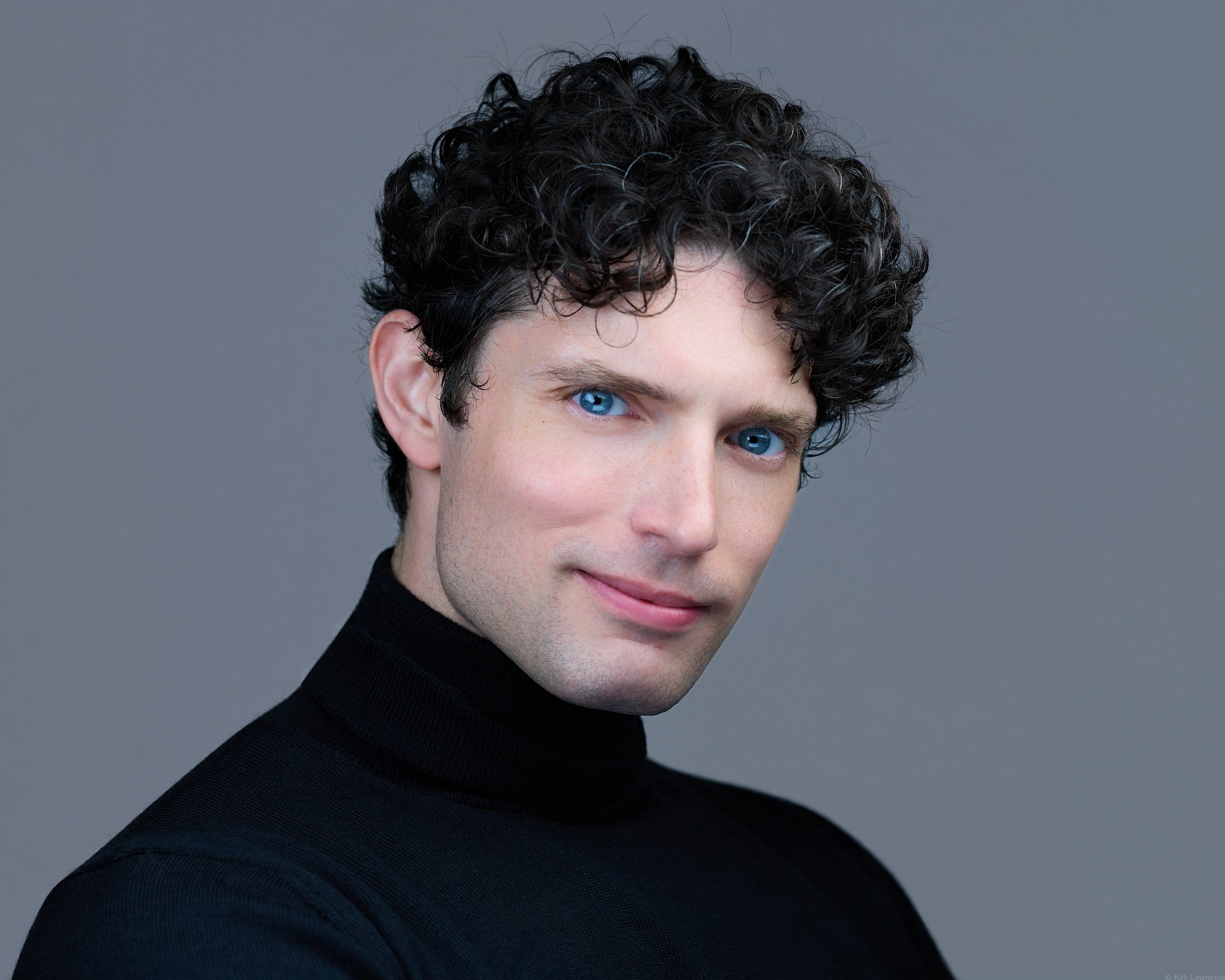 Close-up of a young man with curly black hair and bright blue eyes wearing a black turtleneck against a gray background.