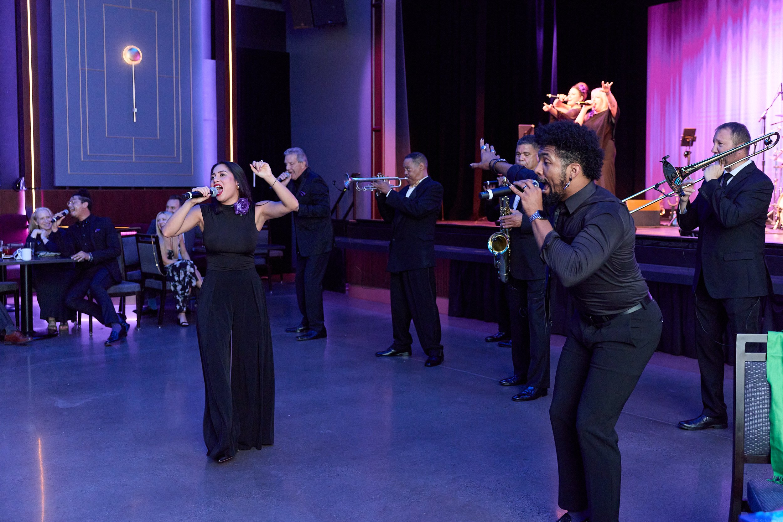 A group of people performing on stage in a nightclub or concert setting, with a female singer and musicians playing instruments. Some audience members are seated at tables, enjoying the performance.