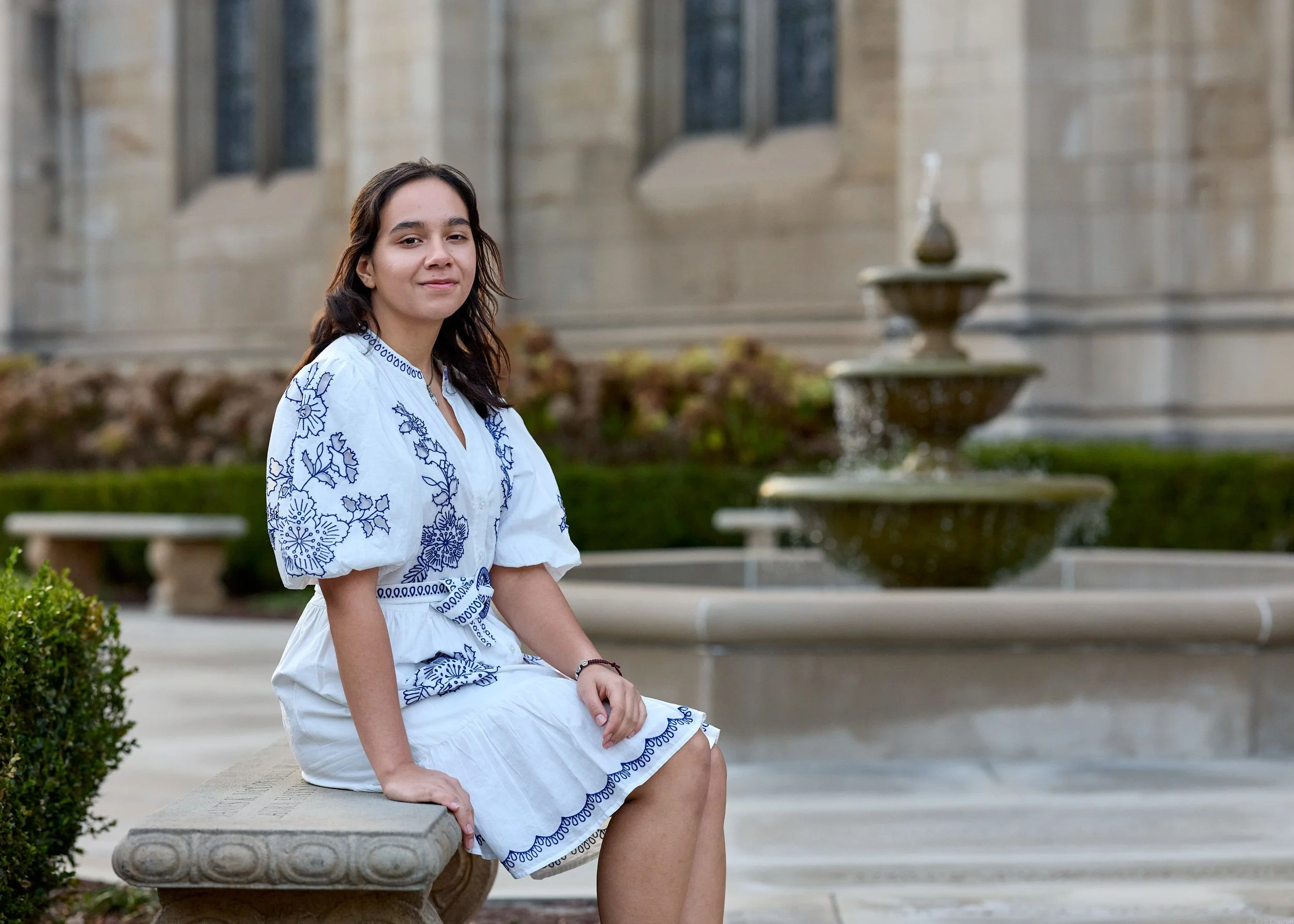 A young woman with dark hair and light skin sitting on a stone bench outdoors near a fountain, wearing a white dress with blue embroidery.