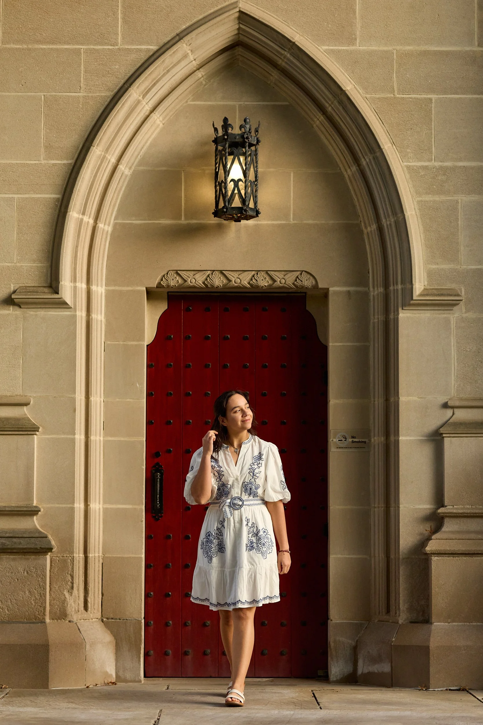 Young woman wearing a white dress with blue embroidery, standing in front of a large red door with black studs under an arched stone entryway, with a decorative lantern above.
