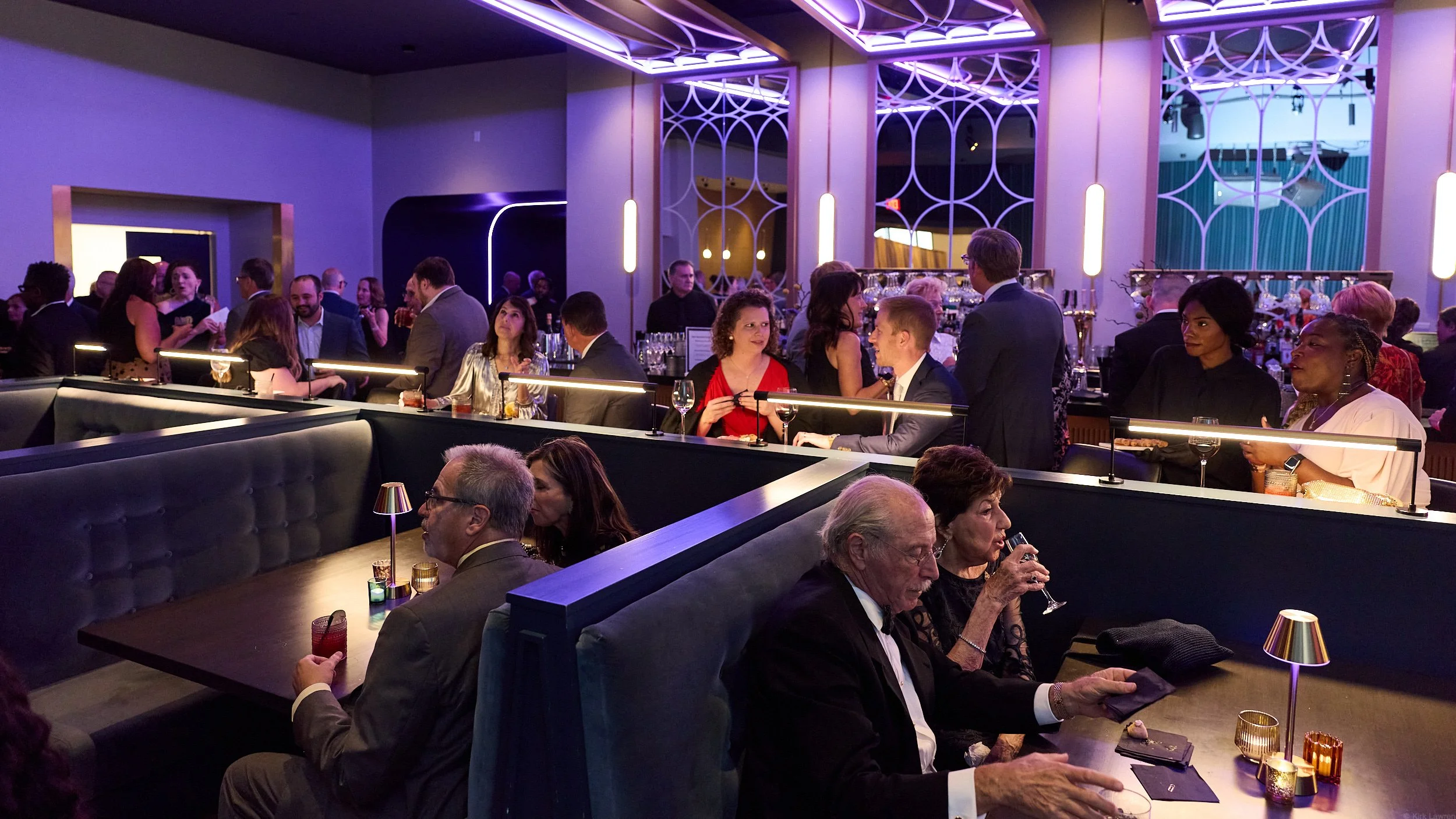 Scene of a sophisticated social gathering or party inside a modern venue with dim, colorful lighting. Guests are seated and mingling, with some standing around a bar area. The atmosphere is lively and elegant, featuring contemporary decor with geomet