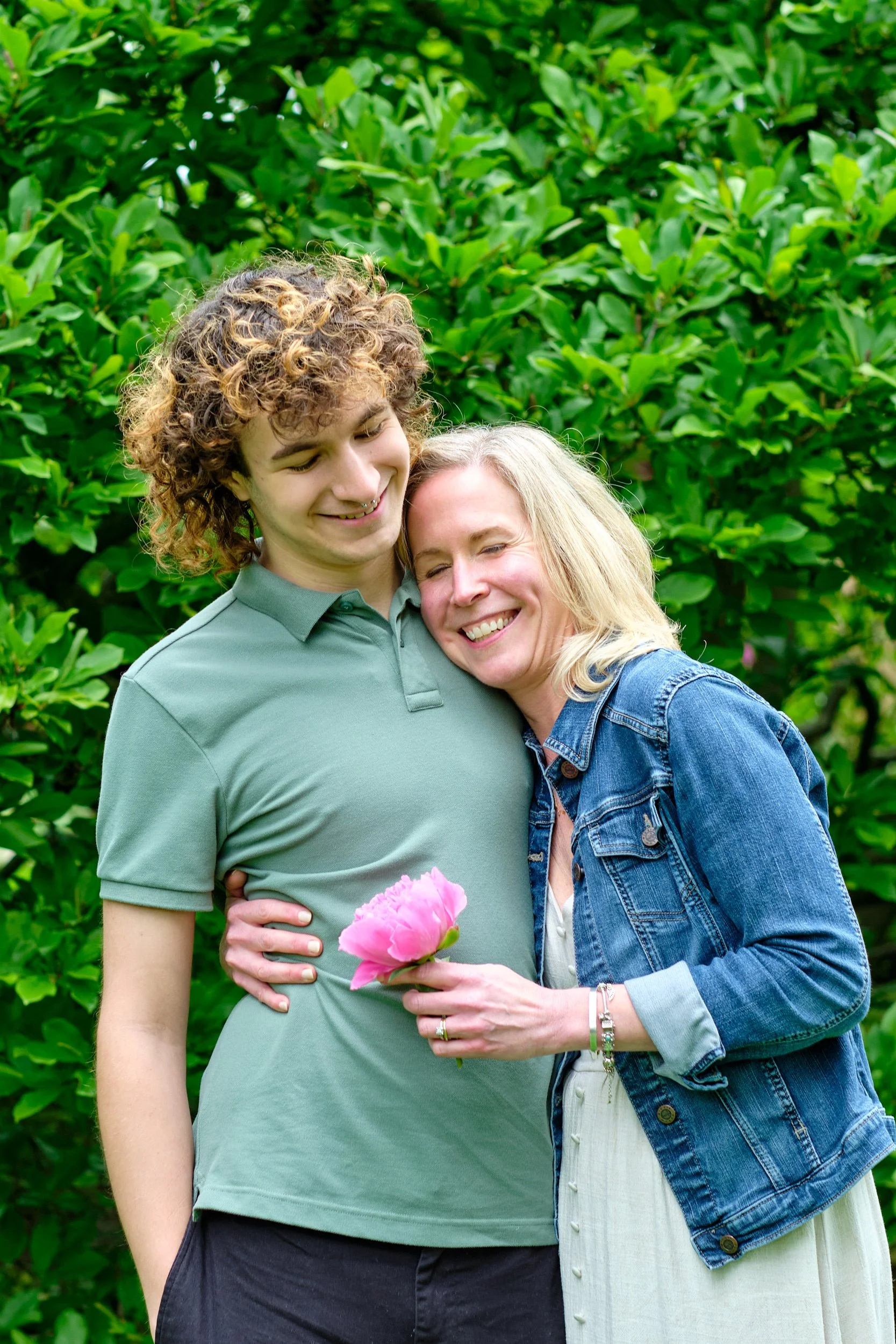 Two women, one with curly blonde hair and the other with straight blonde hair, are hugging outdoors in front of green bushes. The woman with curly hair is holding a pink flower.