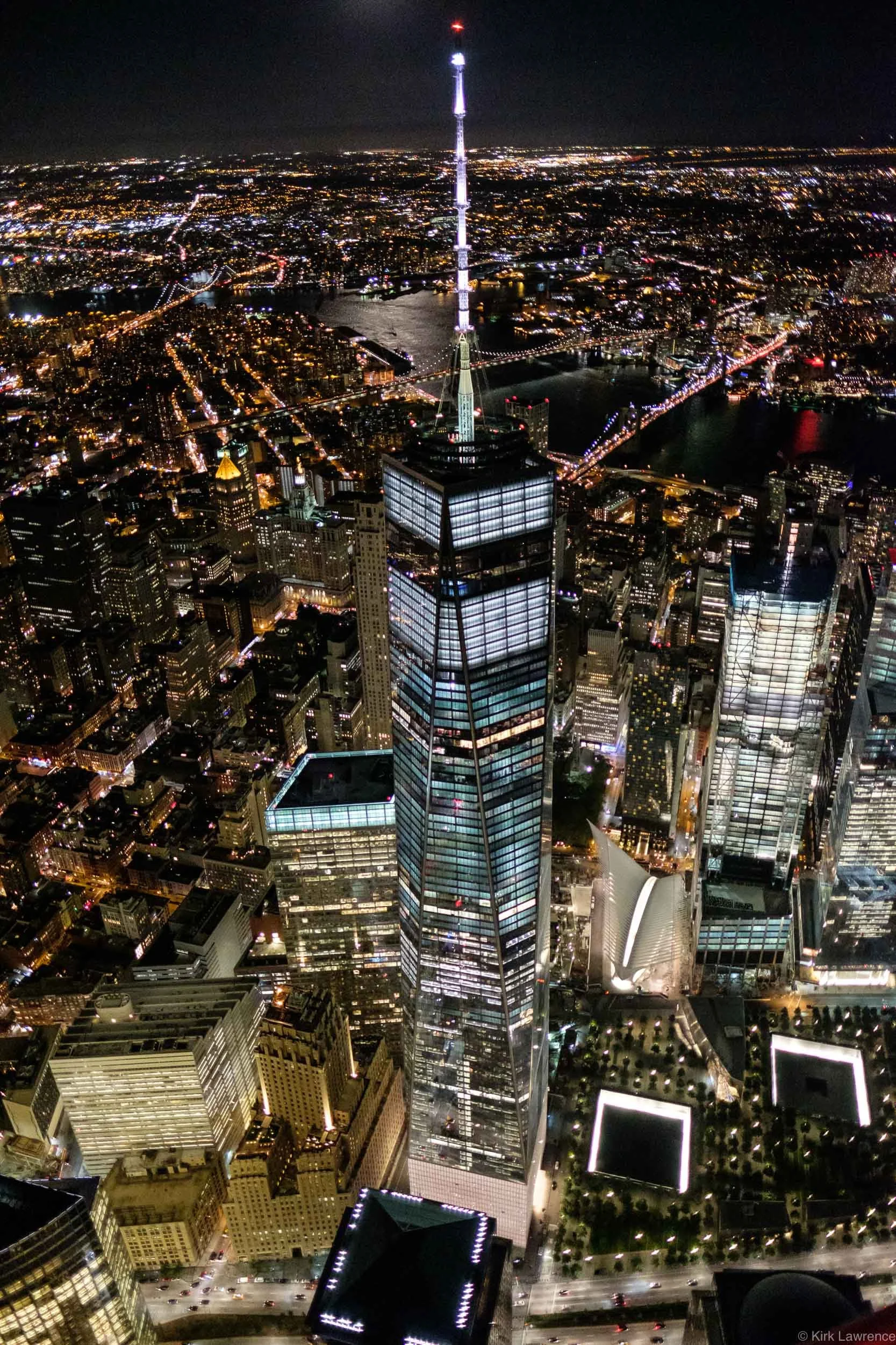 Night view of New York City skyline featuring the illuminated One World Trade Center tower at the center, with surrounding buildings and city lights, and bridges crossing the river.
