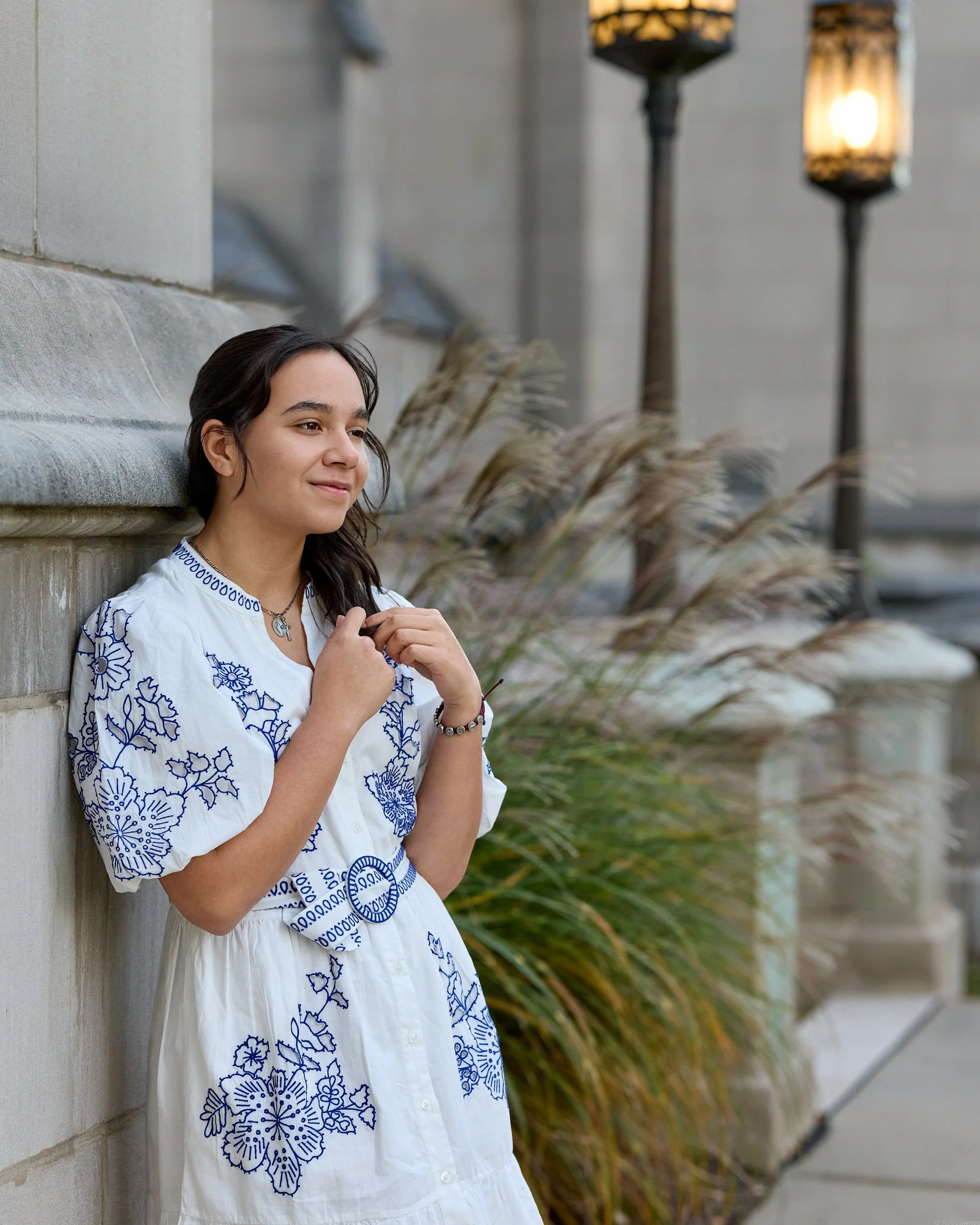 Young woman with dark hair, wearing a white dress with blue floral embroidery, stands against a stone wall, with tall street lamps and ornamental grasses in the background.