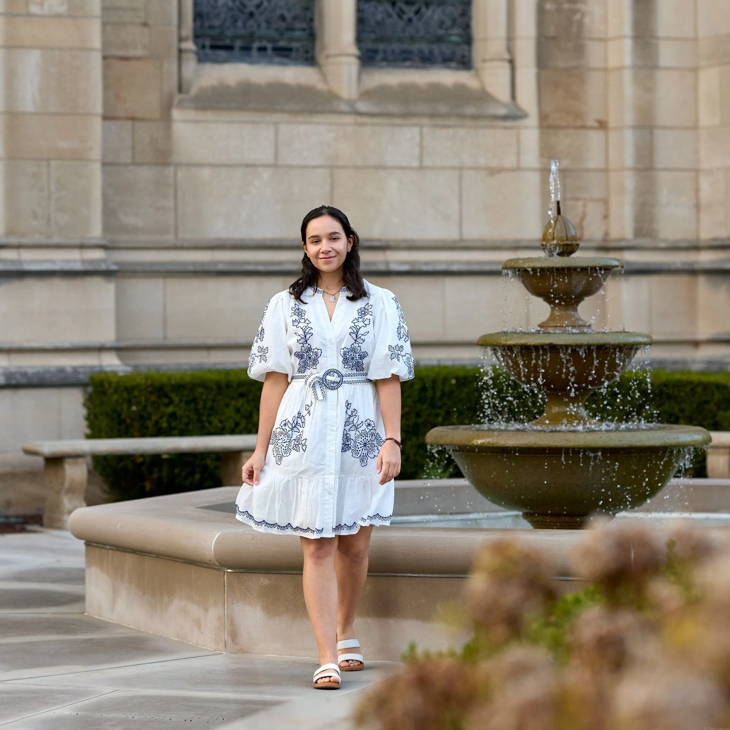 A young woman in a white embroidered dress and white sandals walking near a stone fountain in front of a beige stone building.