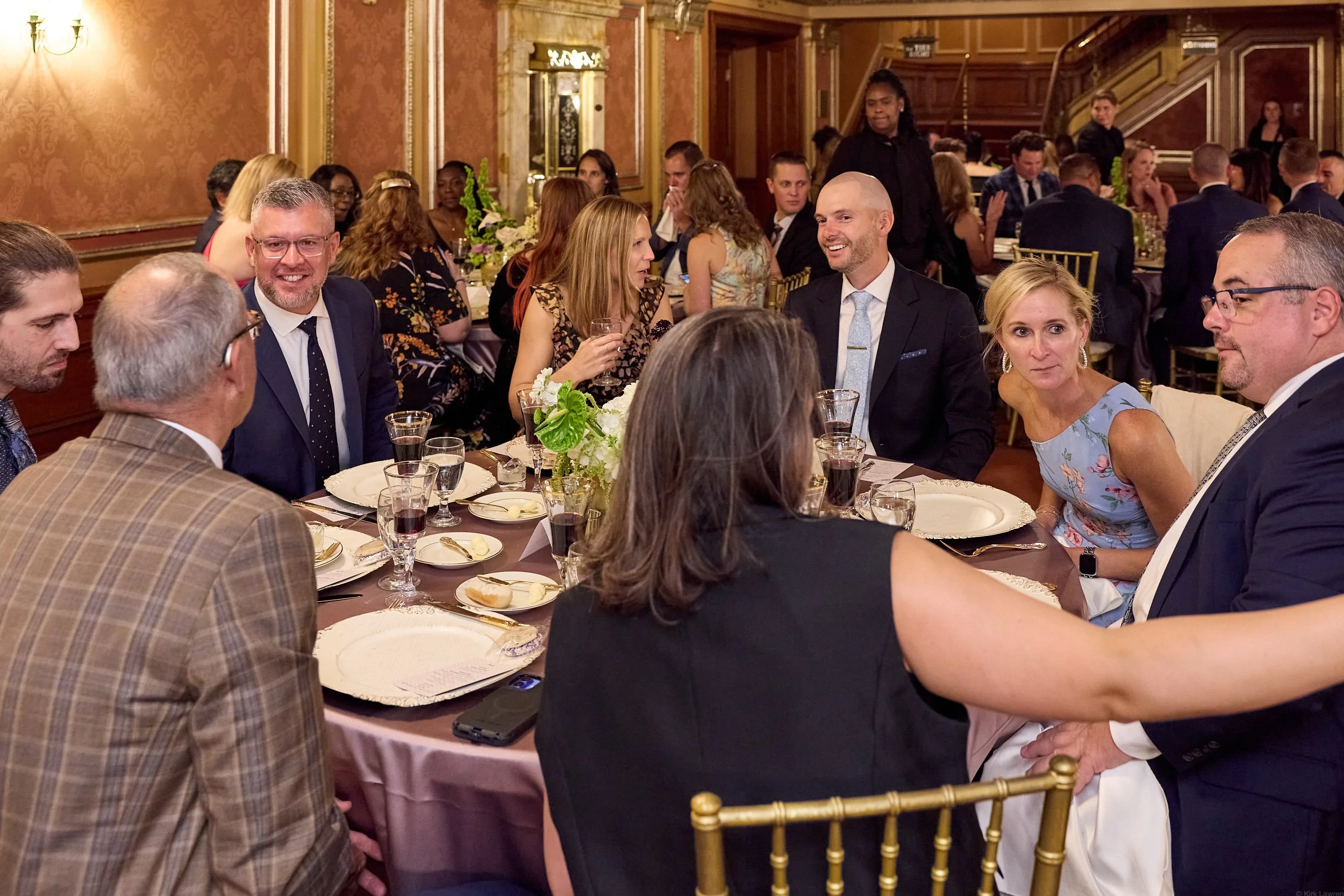 People seated around a dining table at a formal event, engaging in conversation in an ornately decorated room with gold accents and floral centerpieces.