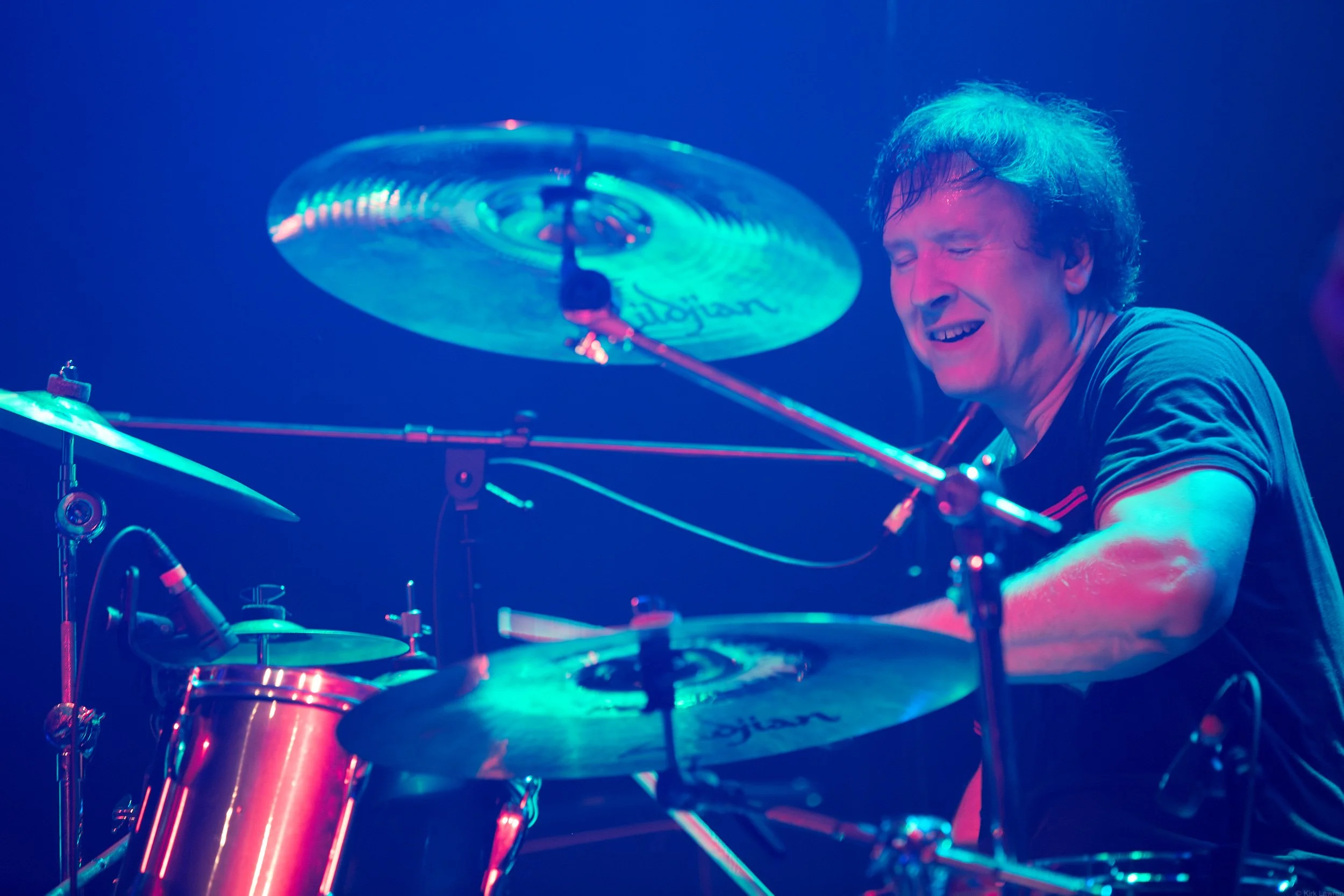 A man playing drums on stage with colorful lighting, smiling with eyes closed.