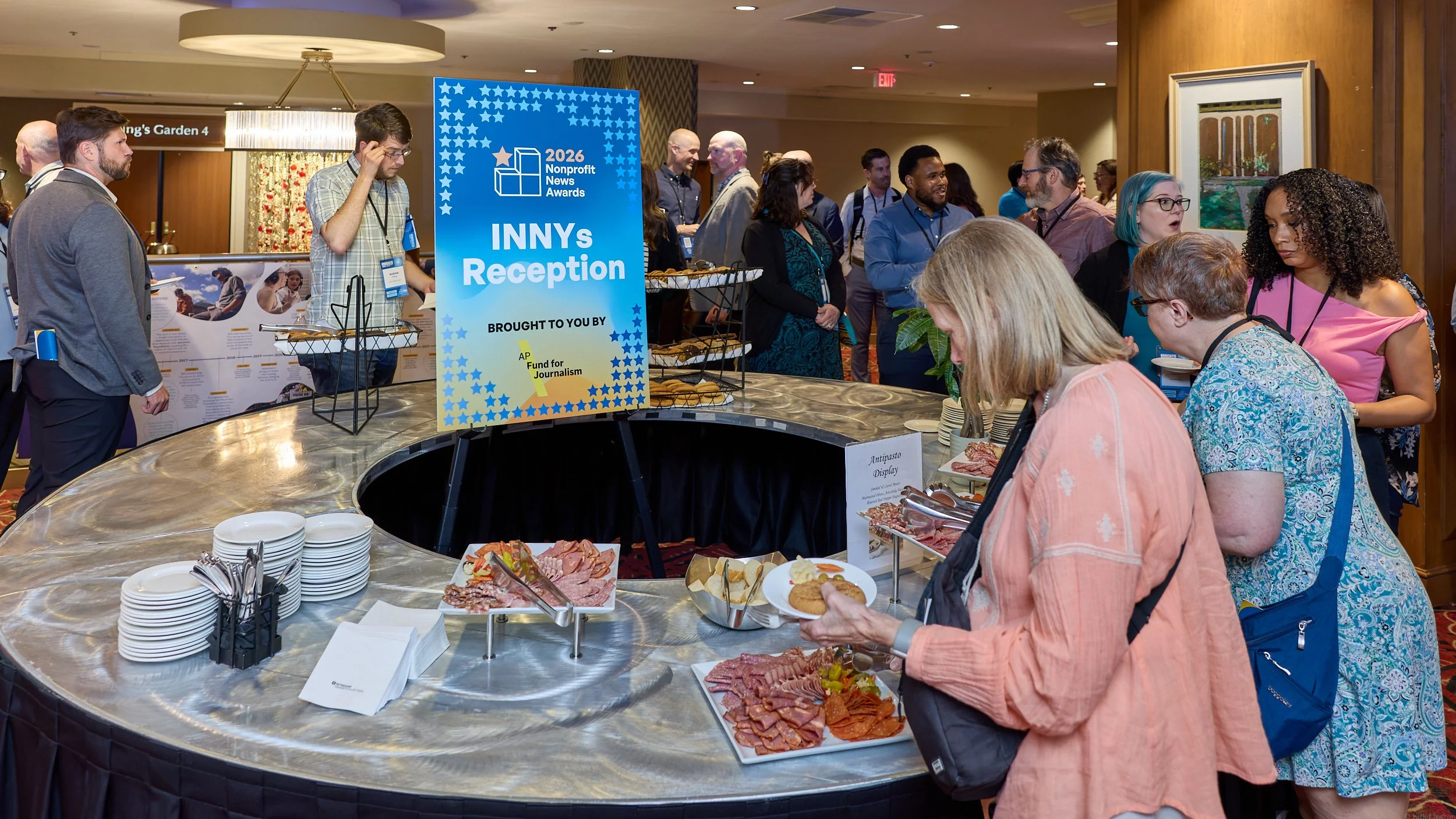People gathering around a buffet at the 2026 Nonprofit News Awards INNYs Reception, with a large blue sign and plates of food on the table.