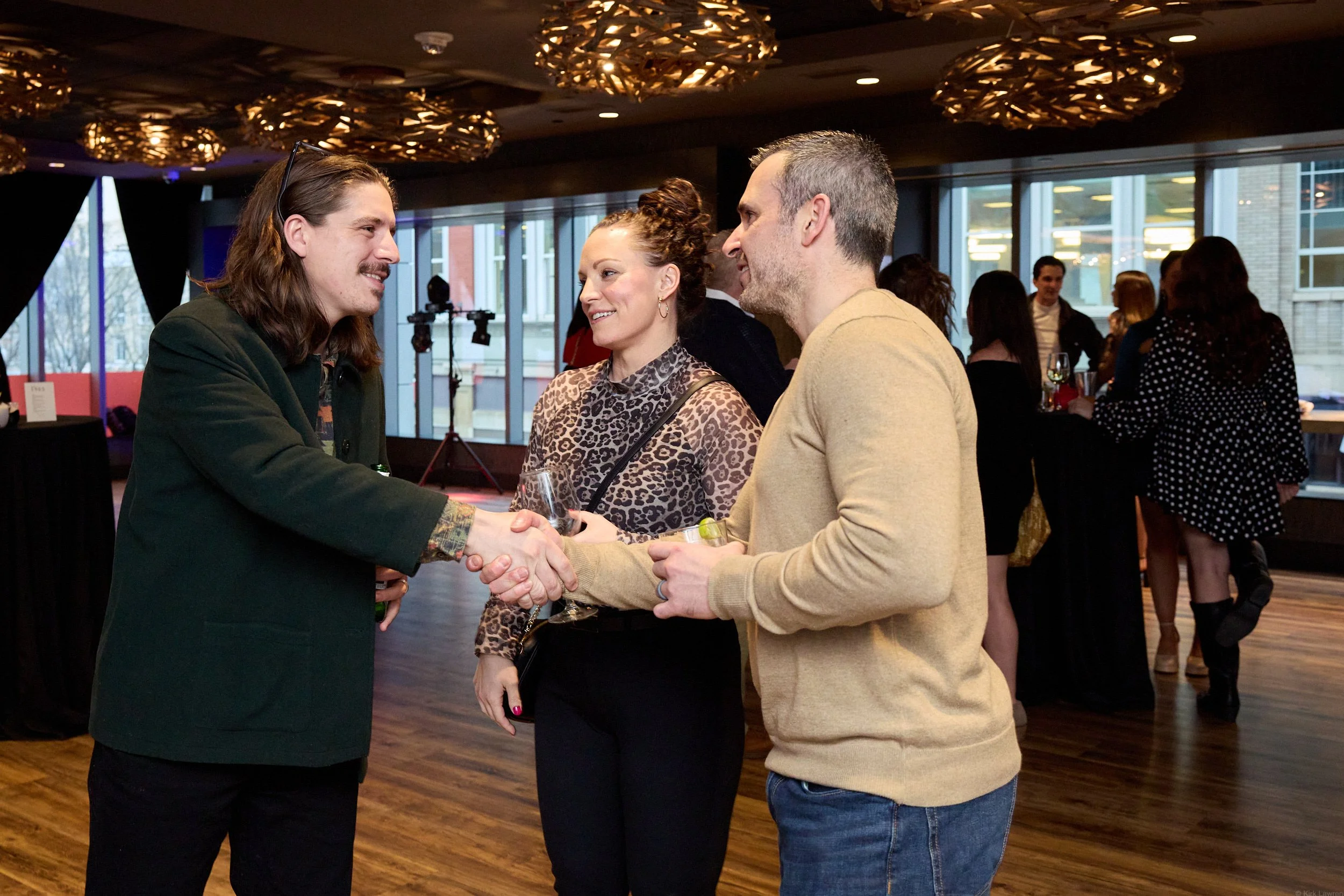Two men shaking hands and smiling at each other while holding glasses of wine, inside a modern event space with other people in the background.