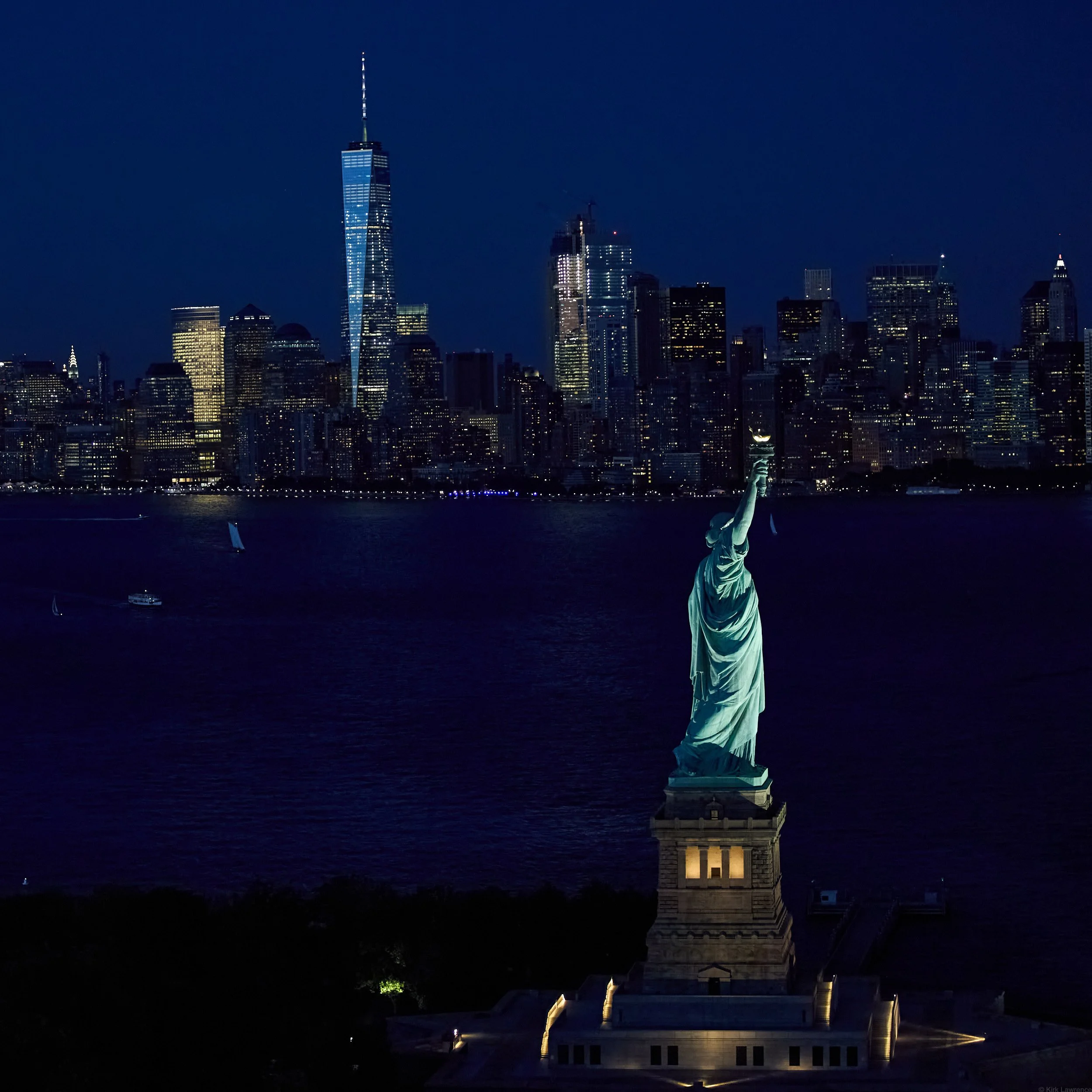 Nighttime view of the Statue of Liberty with illuminated city skyline including the One World Trade Center towering above, on a dark sky.
