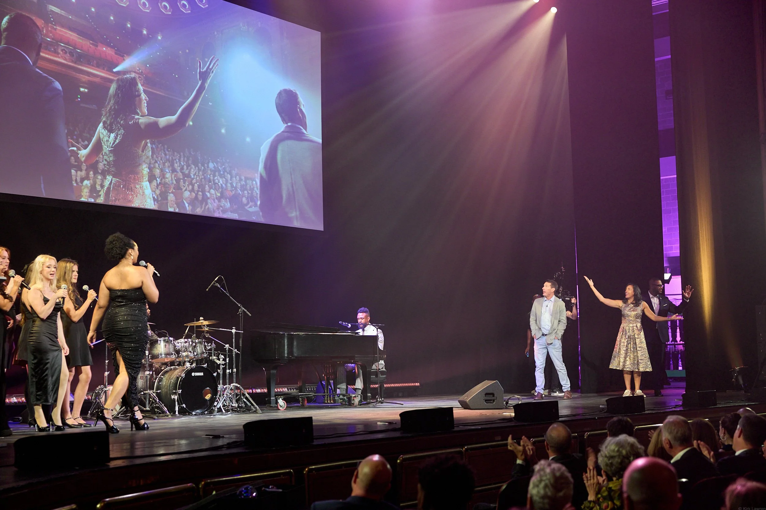 A group of performers on stage during a live concert or award show, with some singers singing into microphones, a pianist playing piano, a drummer behind a drum set, and some individuals dancing or speaking. A large screen above displays an audience 