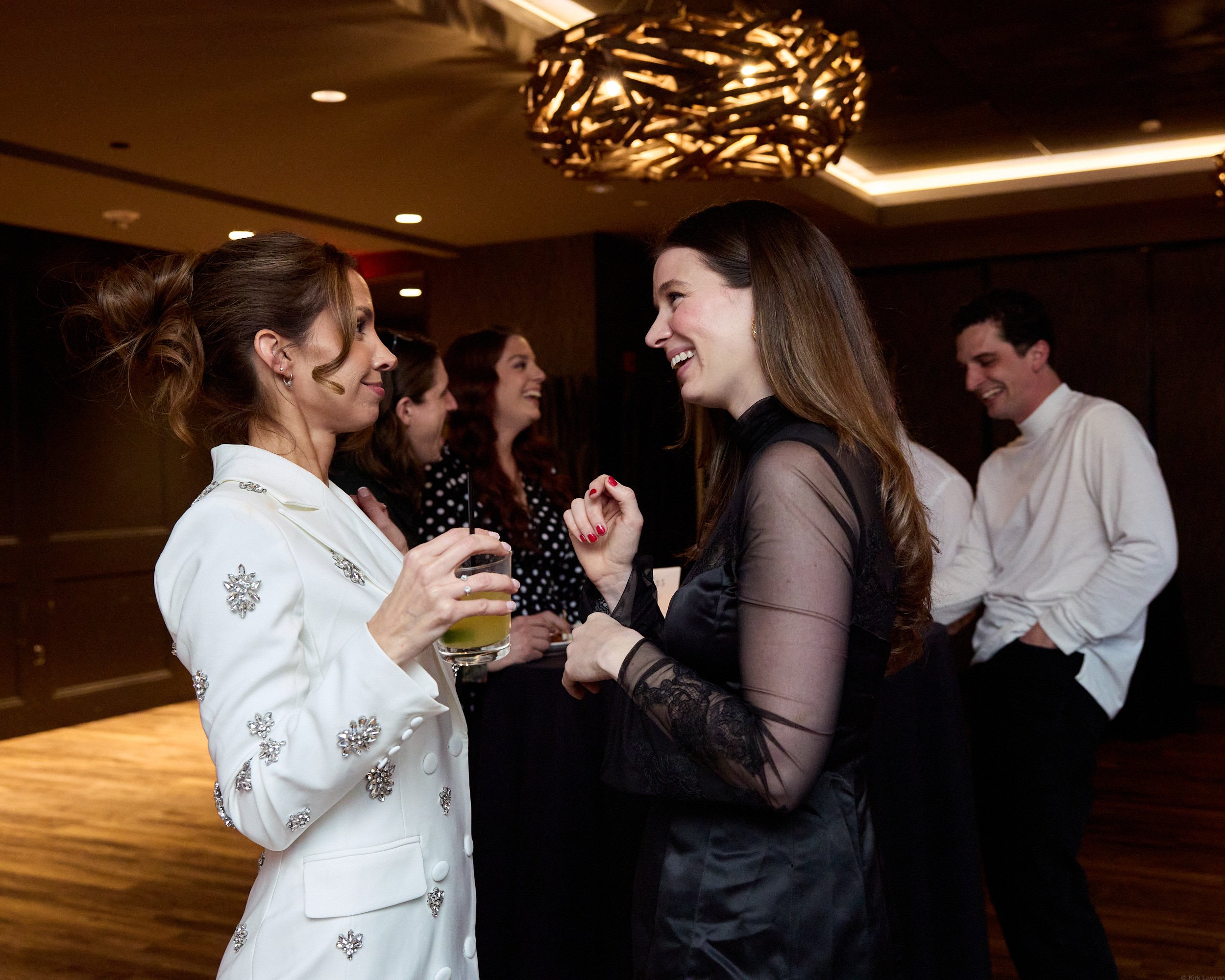 Two women are engaged in a conversation at a social event, smiling and looking into each other's eyes. One woman has curly hair and is wearing a white dress with floral embellishments, holding a drink, while the other woman has straight hair and is w
