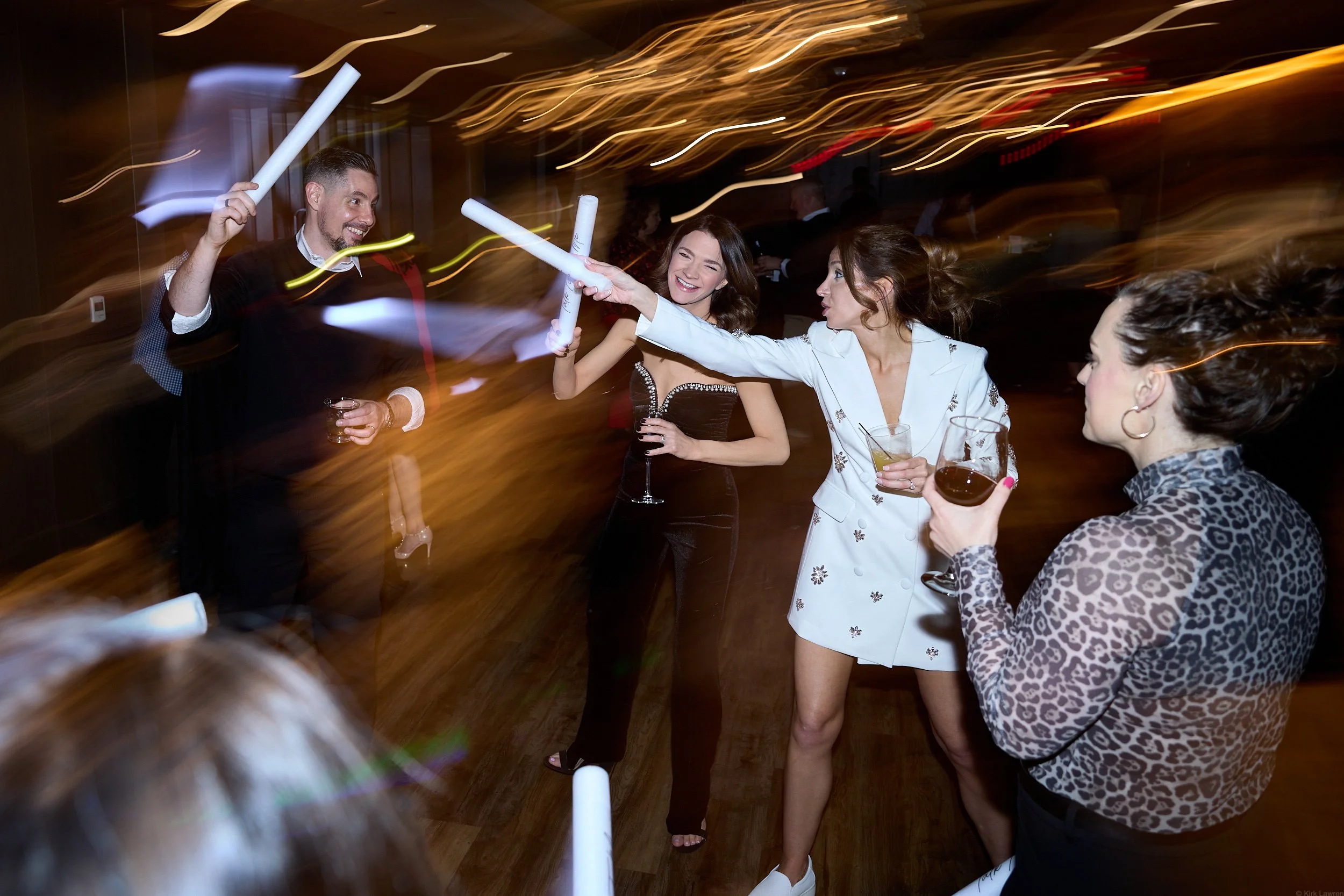 Party scene with five women and one man celebrating, holding foam sticks, in a dimly lit venue with swirling light trails.