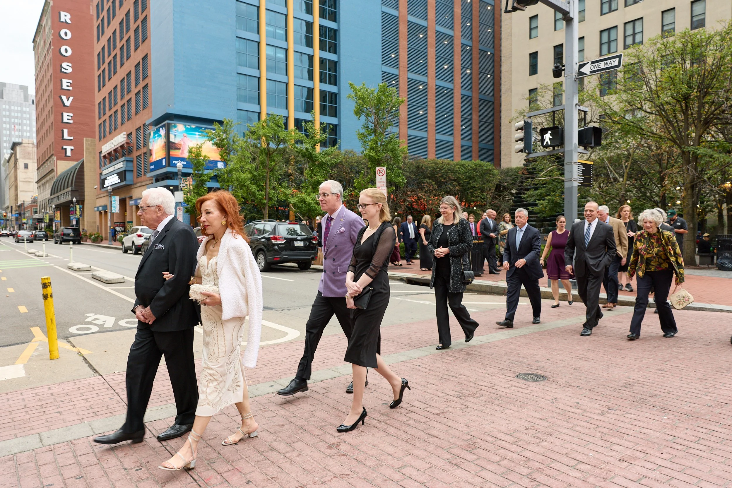 People dressed in business attire crossing a city street in front of high-rise buildings, traffic lights, and trees on a partly cloudy day.