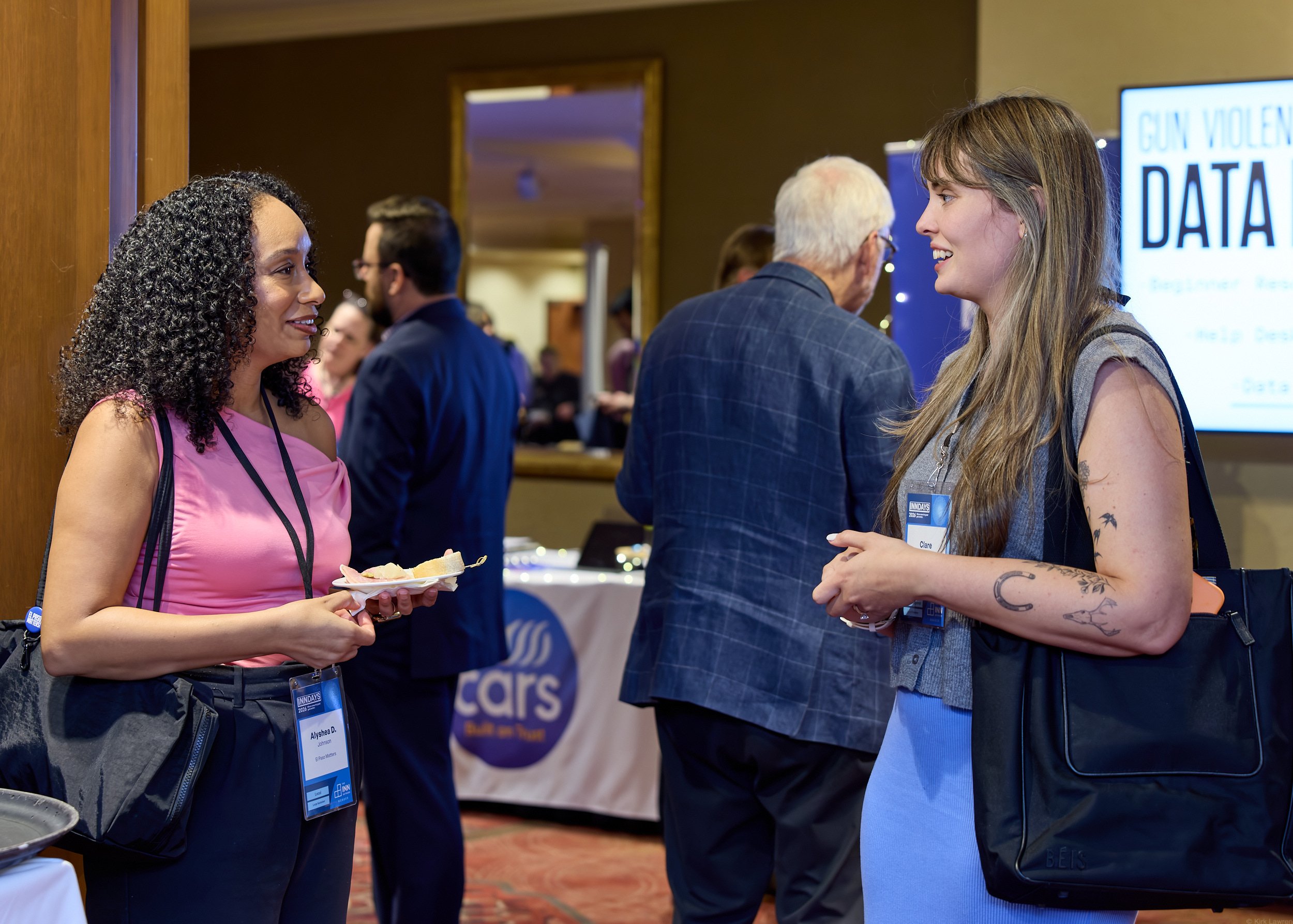Two women talking at an indoor event, wearing name tags, with one holding a plate of food. There are people and informational displays in the background.