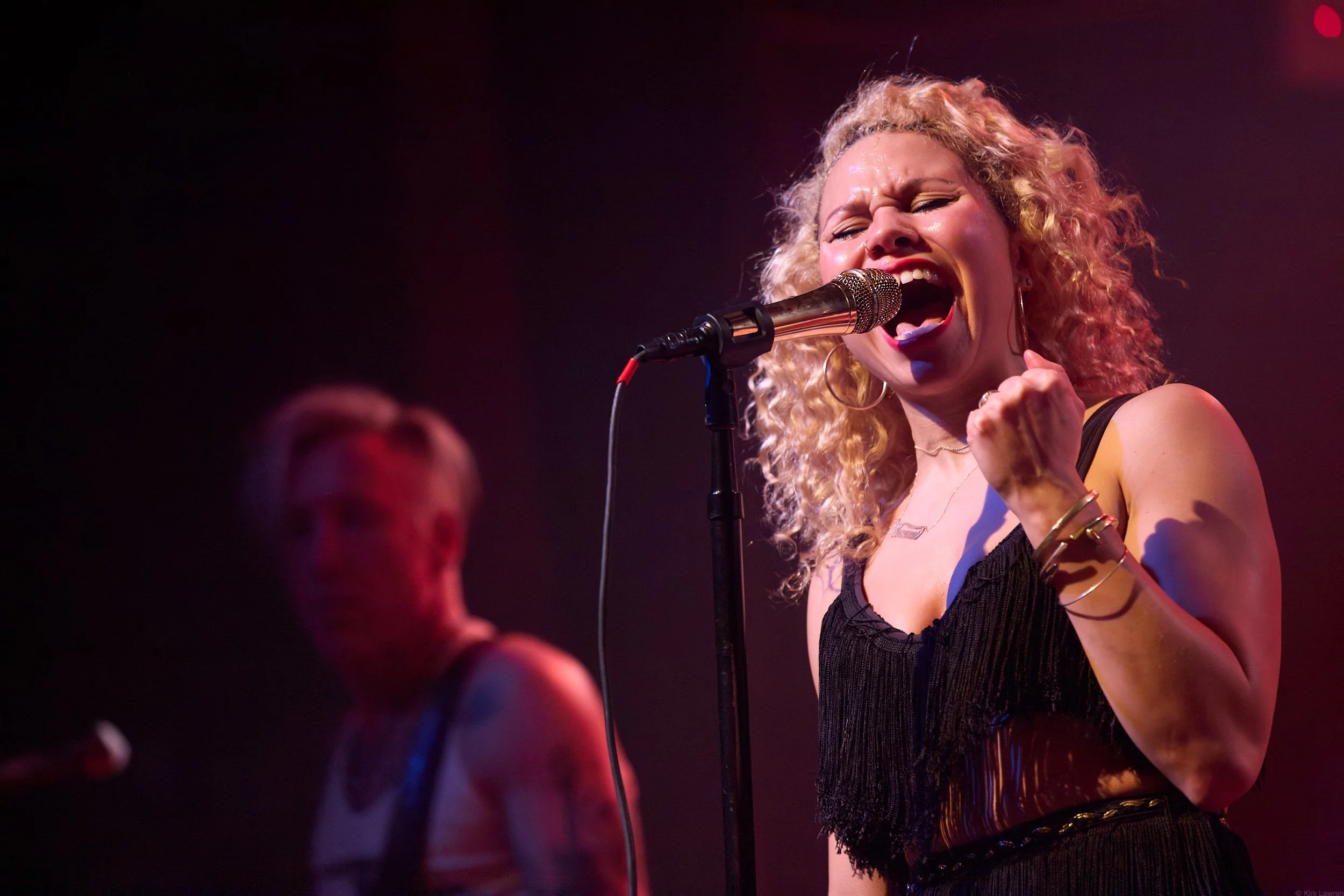 A woman singing passionately into a microphone on stage during a performance, with an expressive facial expression and curly blonde hair.