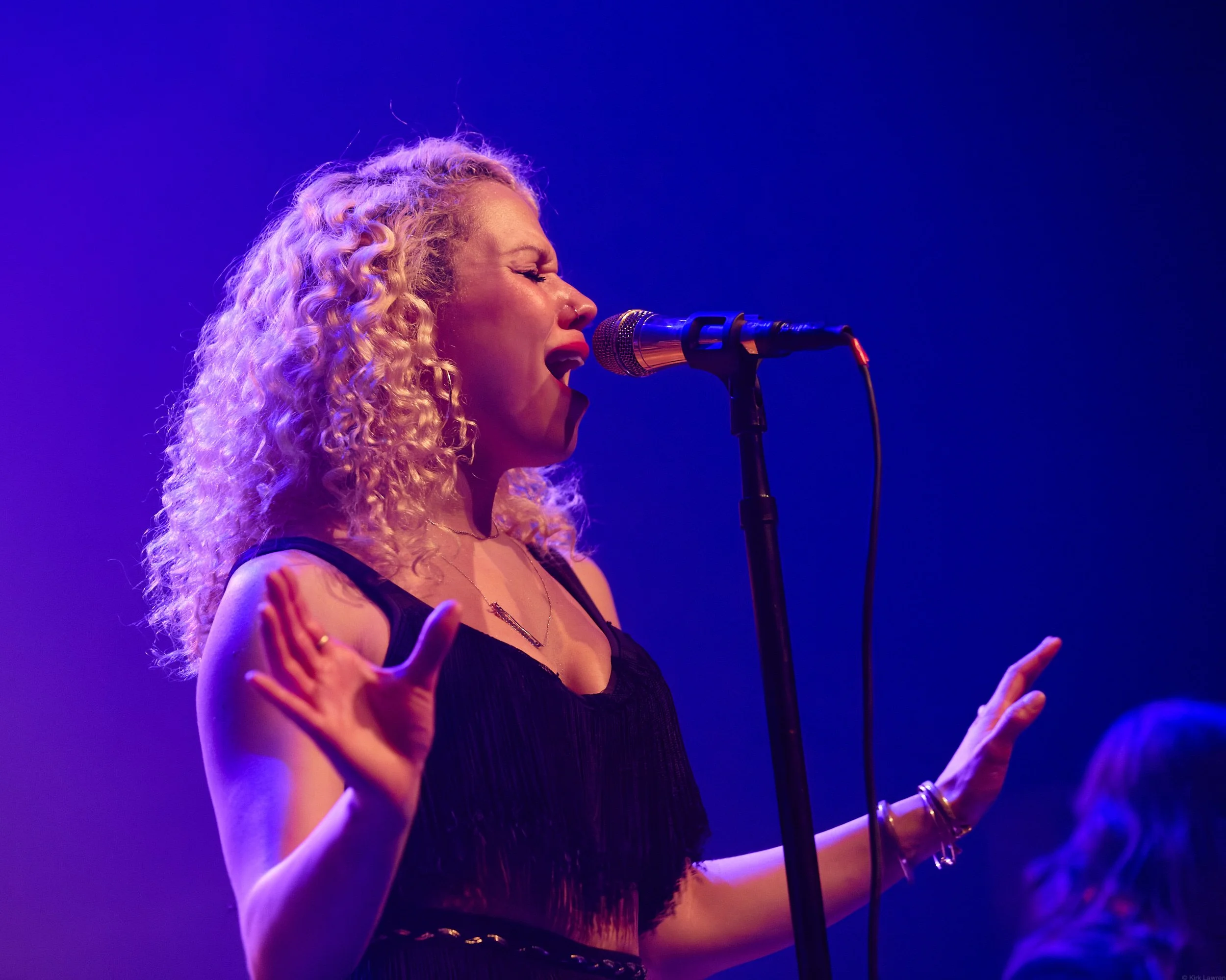 A woman singing into a microphone on stage, with closed eyes and expressive gestures, illuminated by purple and blue stage lights.