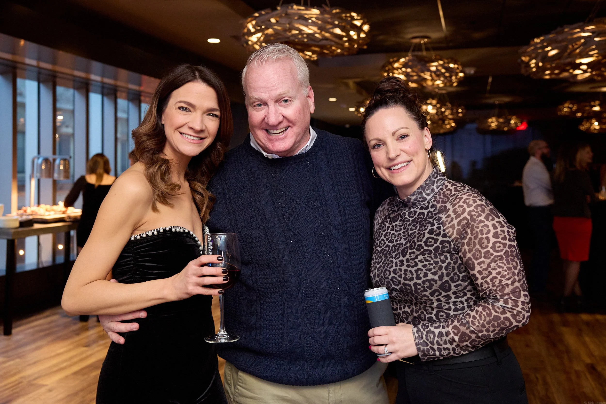 Three people smiling and posing together at an indoor event, one woman holding a glass of red wine, background shows a buffet and other guests at the event.