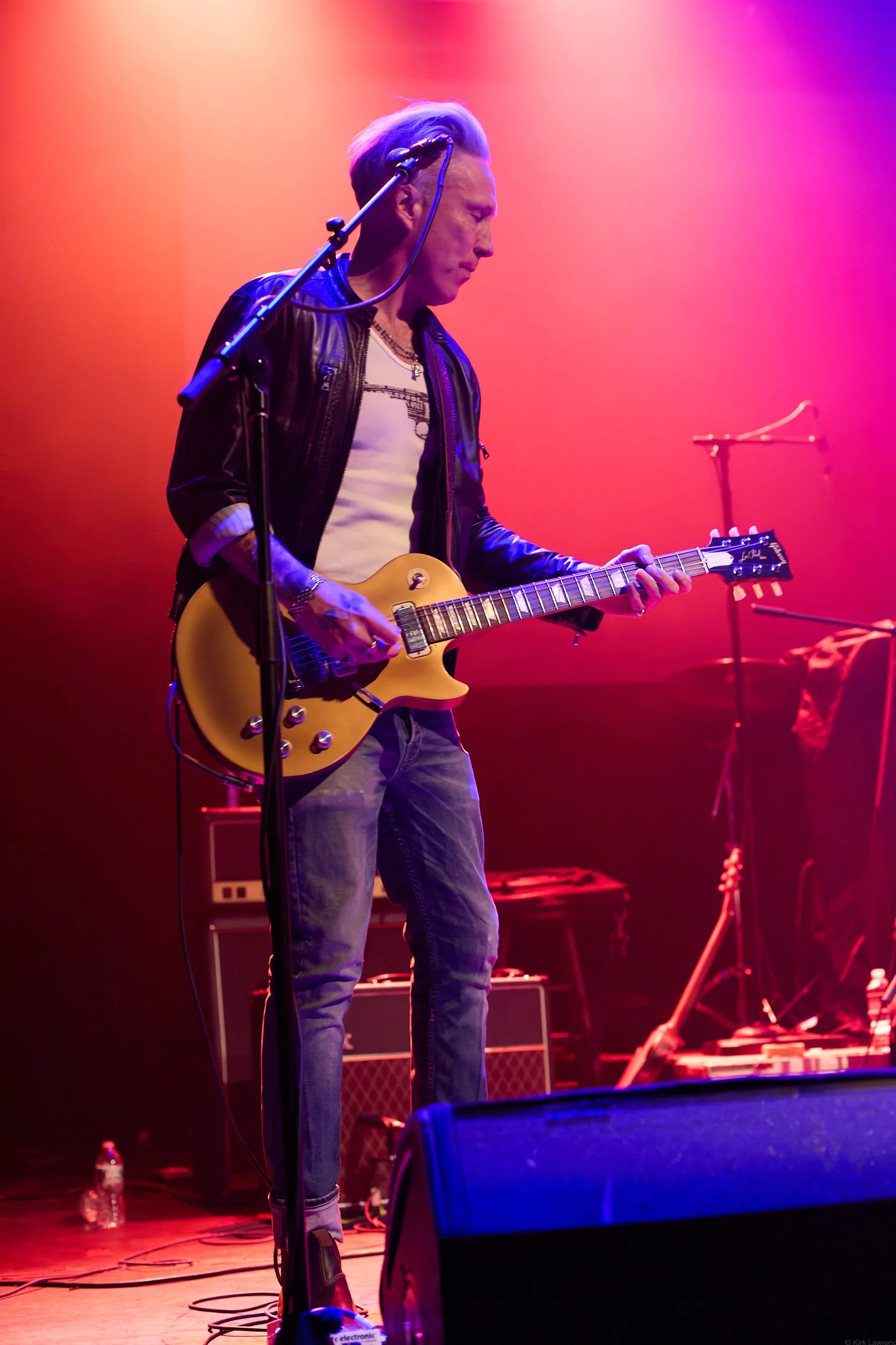 A man with blonde hair, wearing a leather jacket and jeans, playing an electric guitar on stage with colorful purple, pink, and orange lighting.