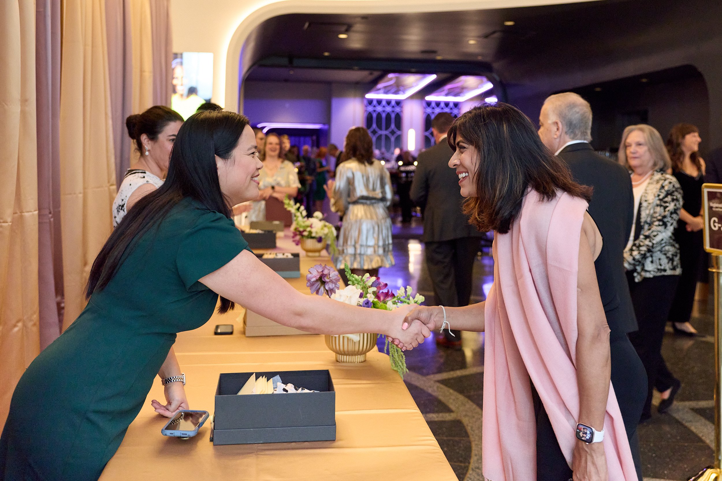 Two women shaking hands at a reception desk in a hotel lobby, smiling and engaging in a friendly conversation.