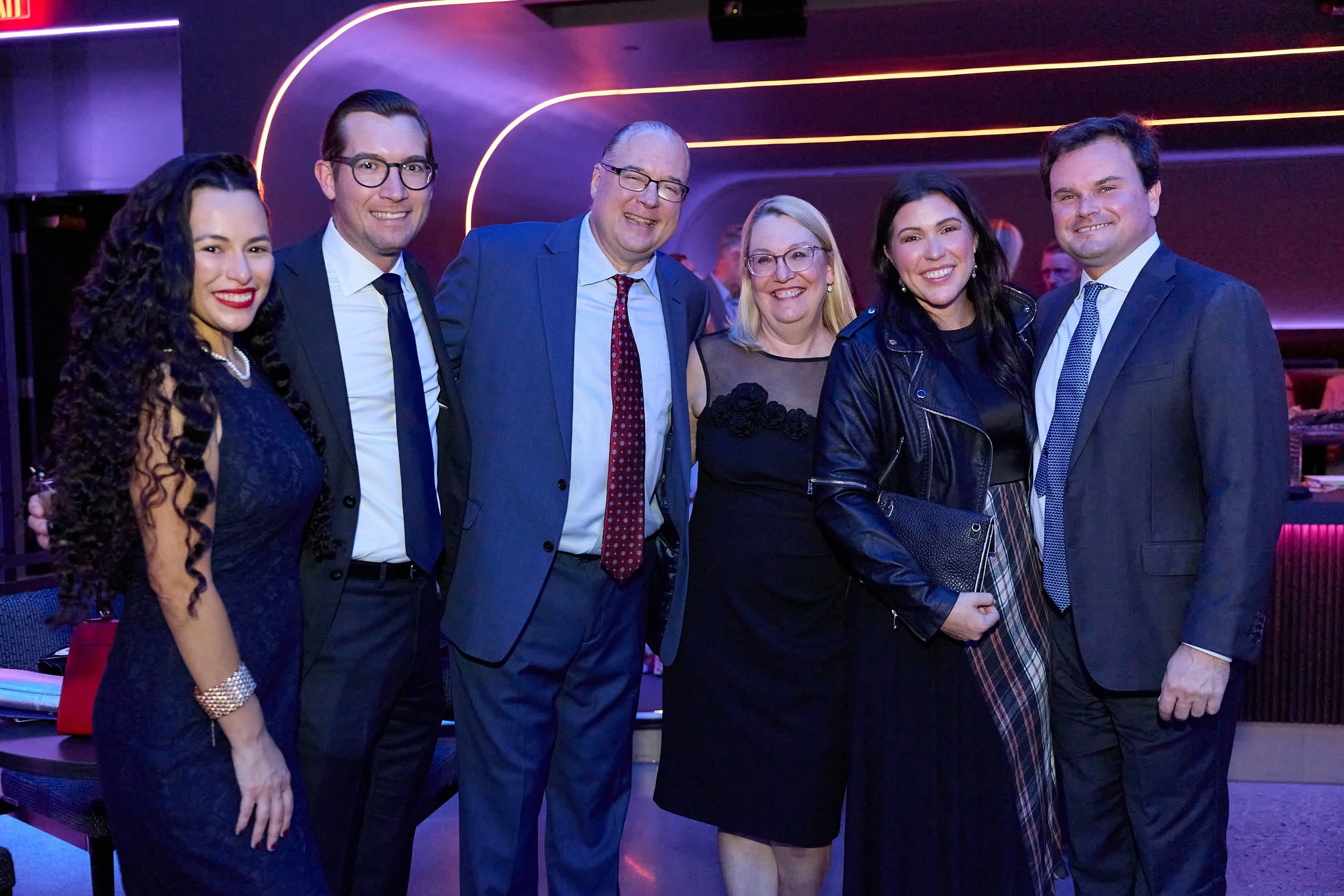 Group of six people dressed in formal attire smiling at a social event with purple and pink lighting.