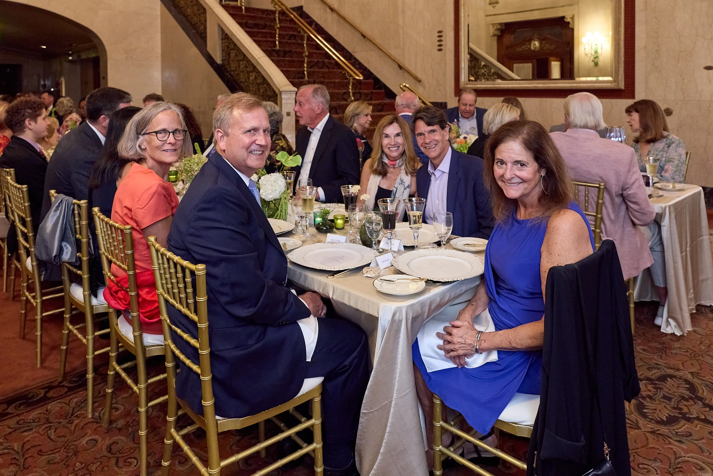 A group of well-dressed people sitting at a banquet table during a formal event in an elegant room with marble walls, a staircase, and decorative lighting.