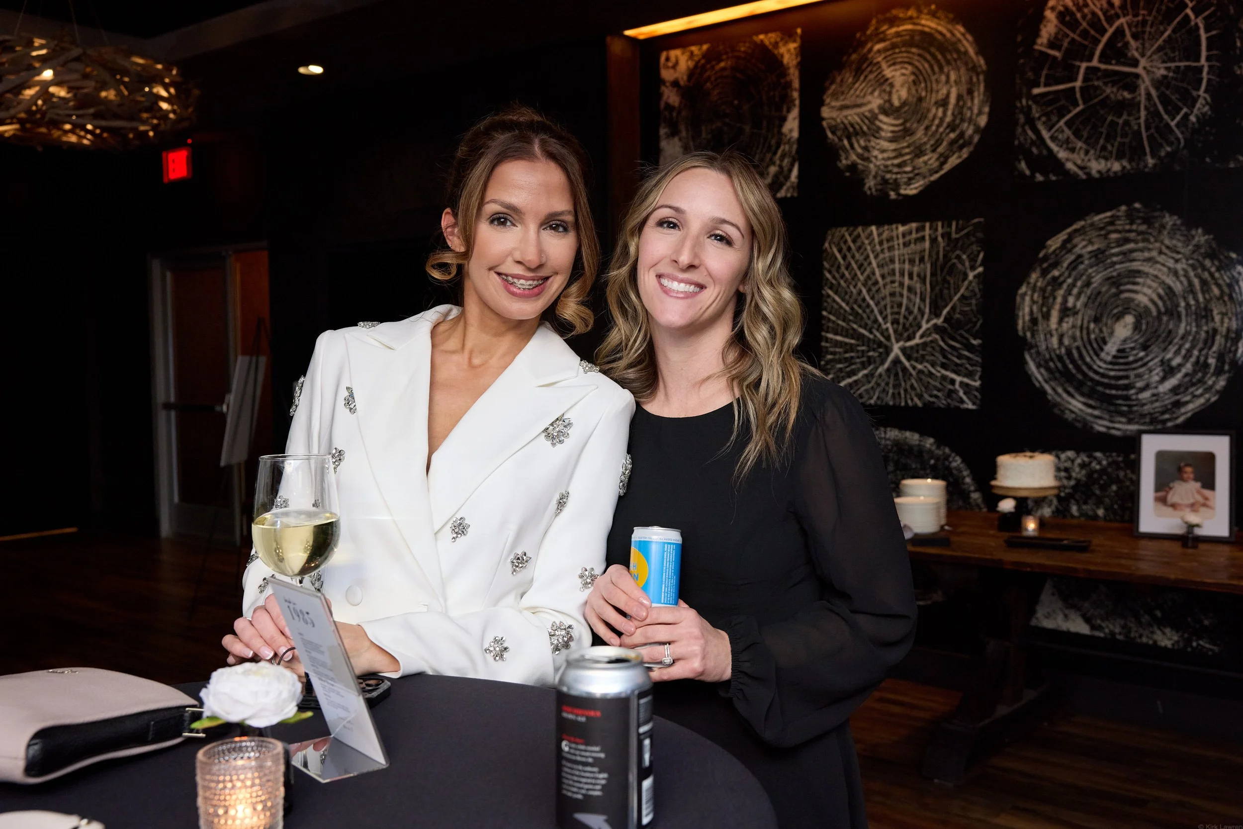 Two women smiling at an indoor event, one holding a wine glass and the other holding a can. They are dressed semi-formally, with a dark, patterned wall in the background. The table in front has a candle and a bouquet of white flowers.