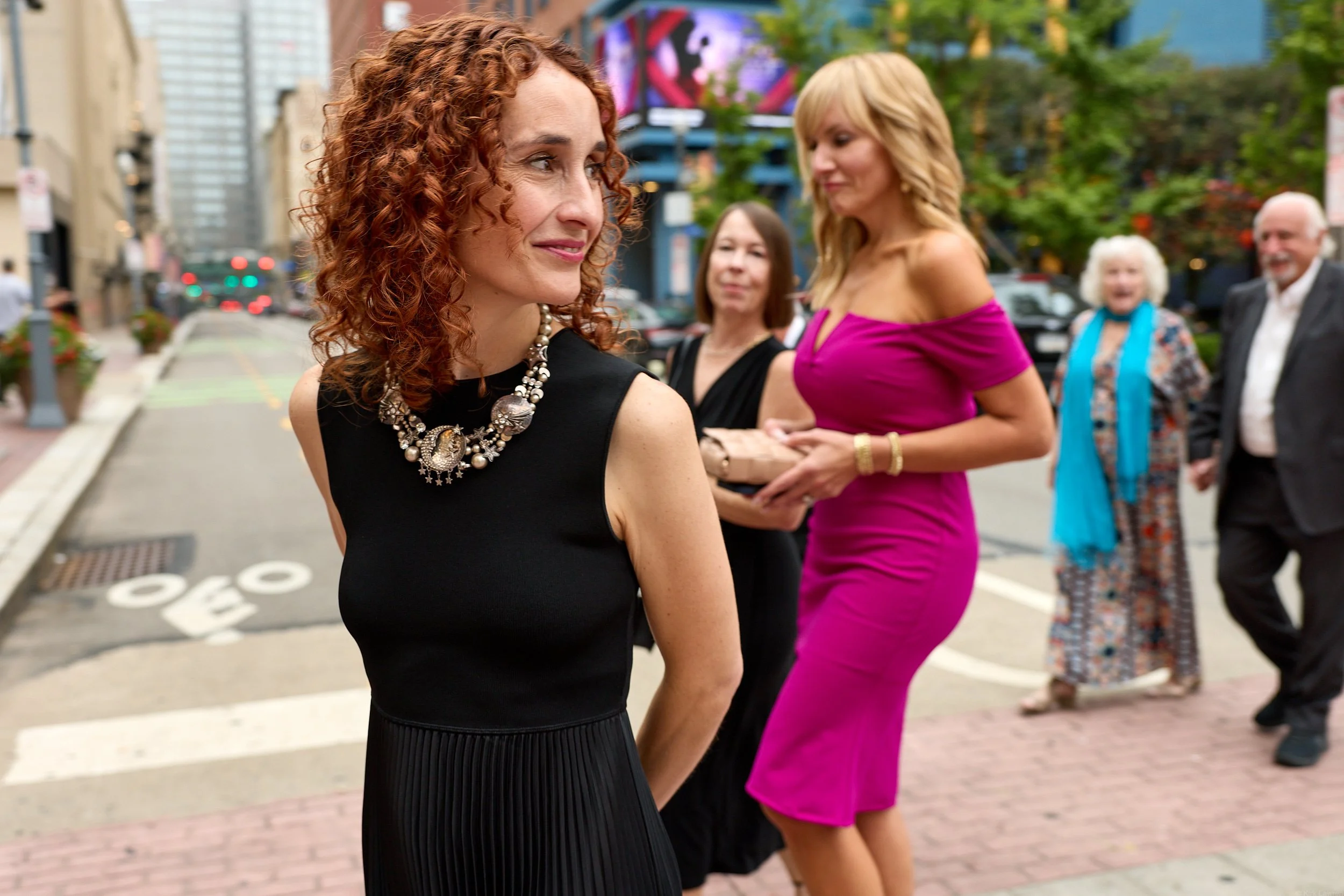 A woman with curly red hair and a black dress with a large pearl and brooch necklace, smiling and looking to her right, in an urban street scene with other women and a man in the background.