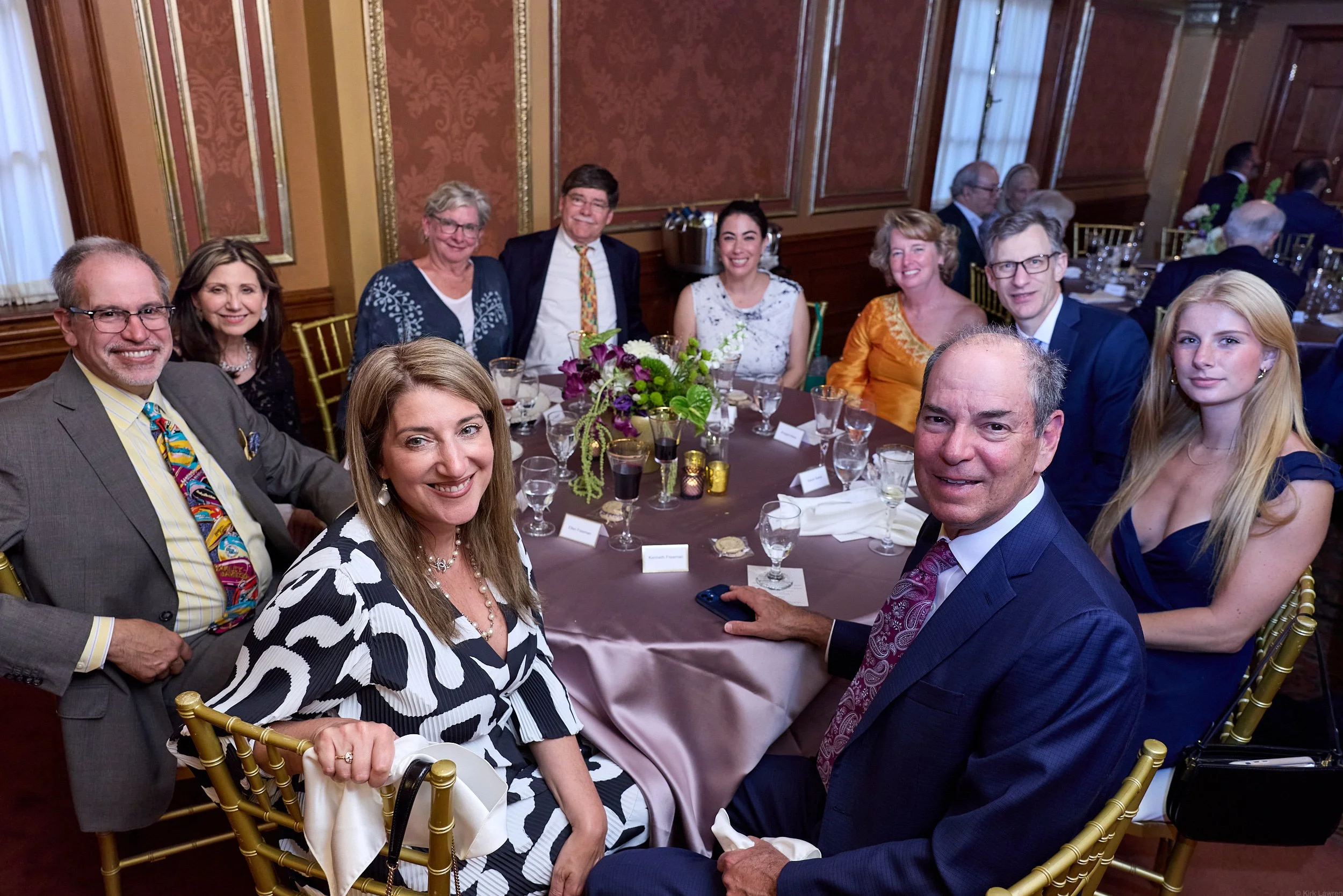 A group of diverse smiling people sitting around a table at a formal event in a decorated room with gold chairs and floral centerpiece.