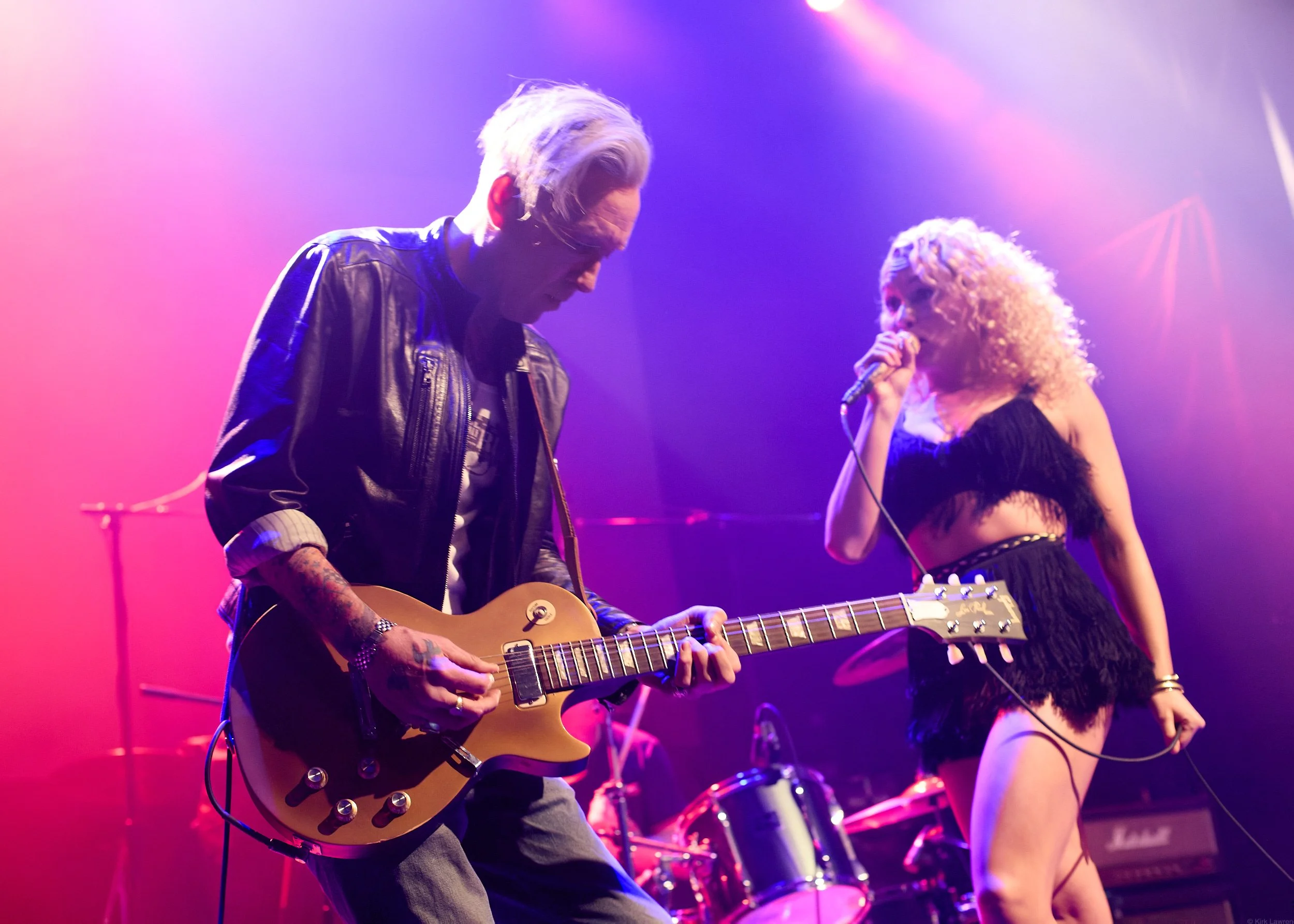 A man playing an electric guitar with a woman singing into a microphone on stage during a live music performance. Colorful stage lights illuminate the scene.