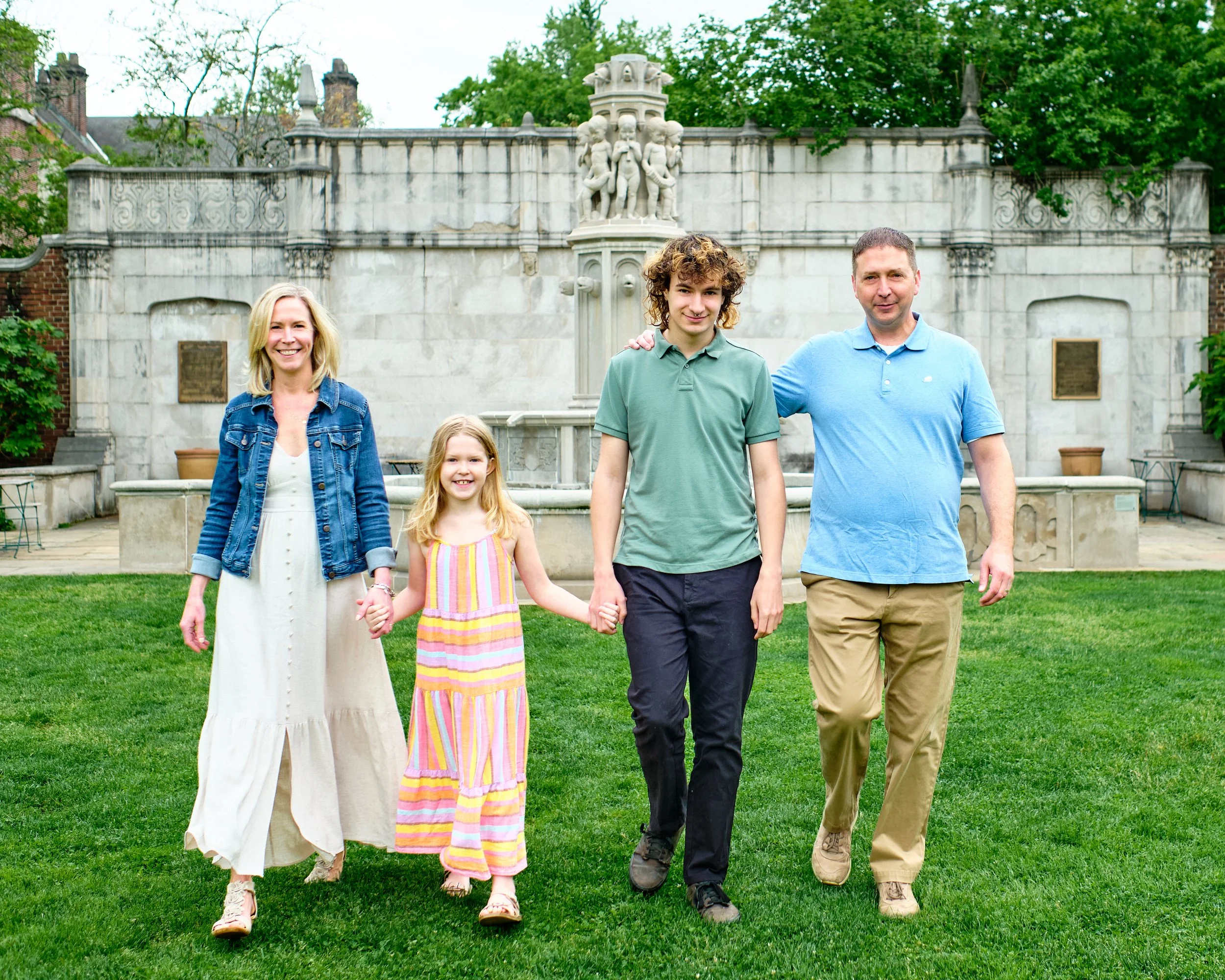 A family of five with three children, two girls and one boy, walking on a green lawn in front of a stone fountain with classical sculptures, trees, and cloudy sky in the background.