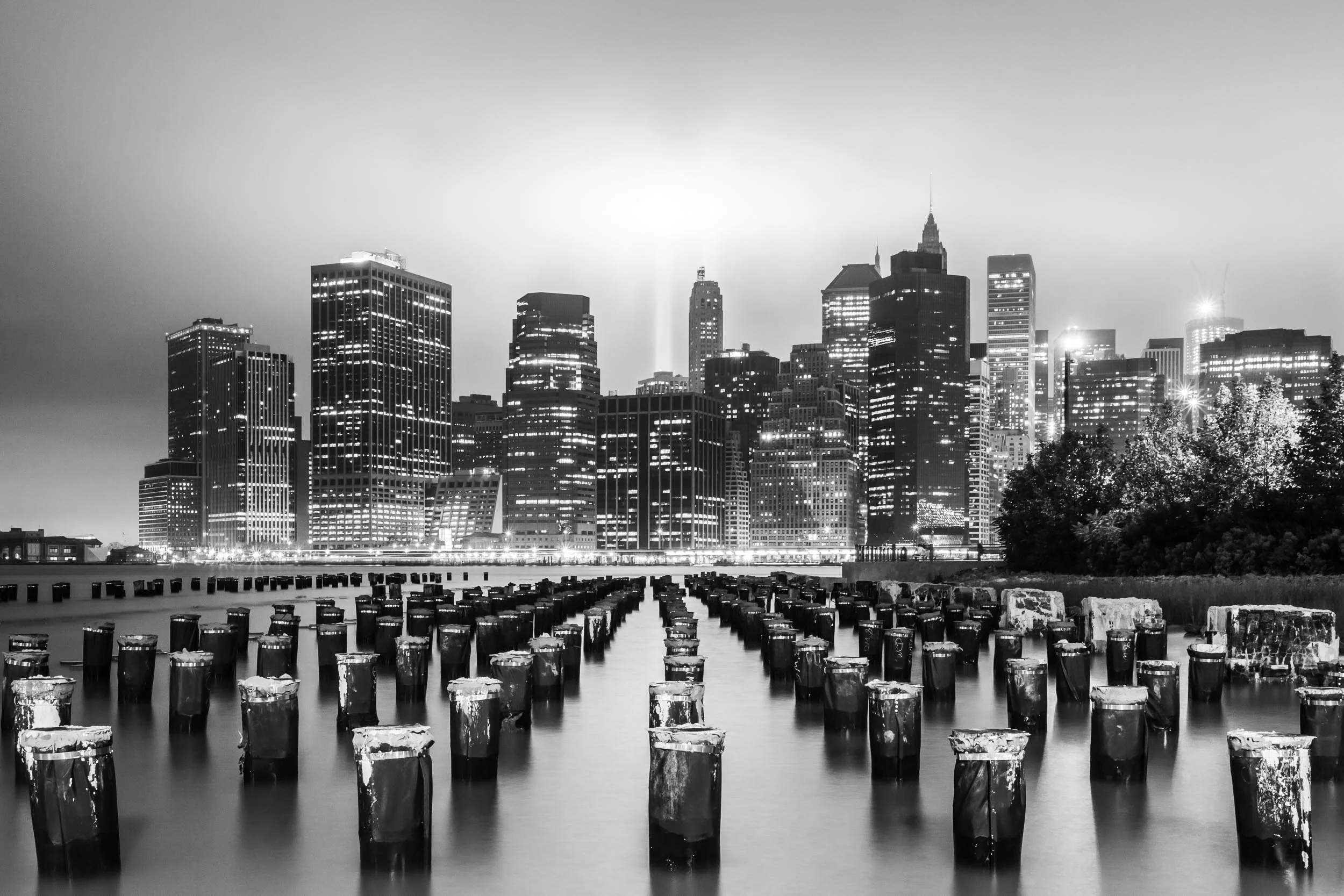 Black and white photo of a Downtown New York City skyline with tall buildings and skyscrapers, viewed from across a body of water with wooden posts in the foreground.