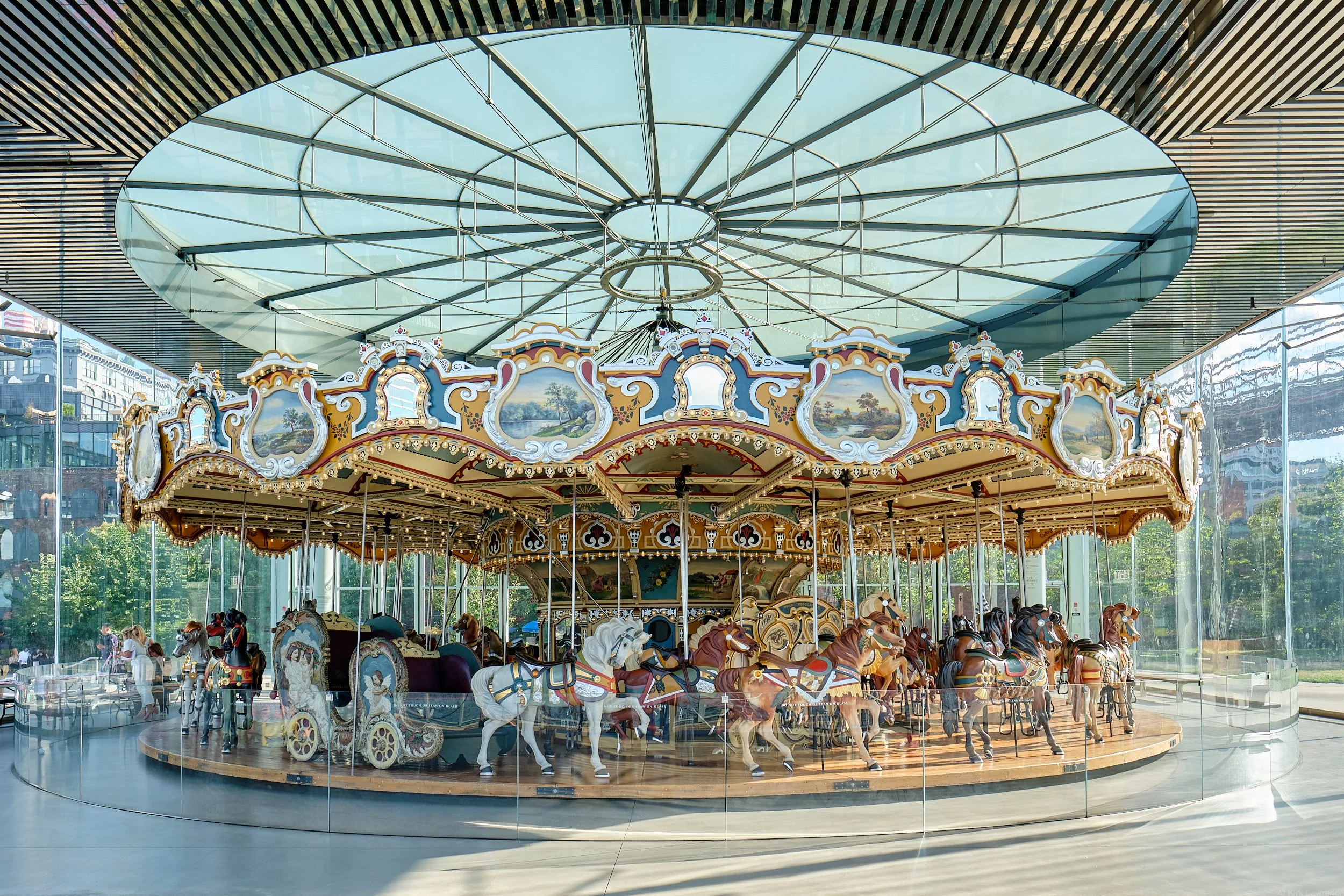 An indoor carousel with decorated horses and carriages, enclosed in a glass and metal structure, with reflections of city buildings on the glass walls.