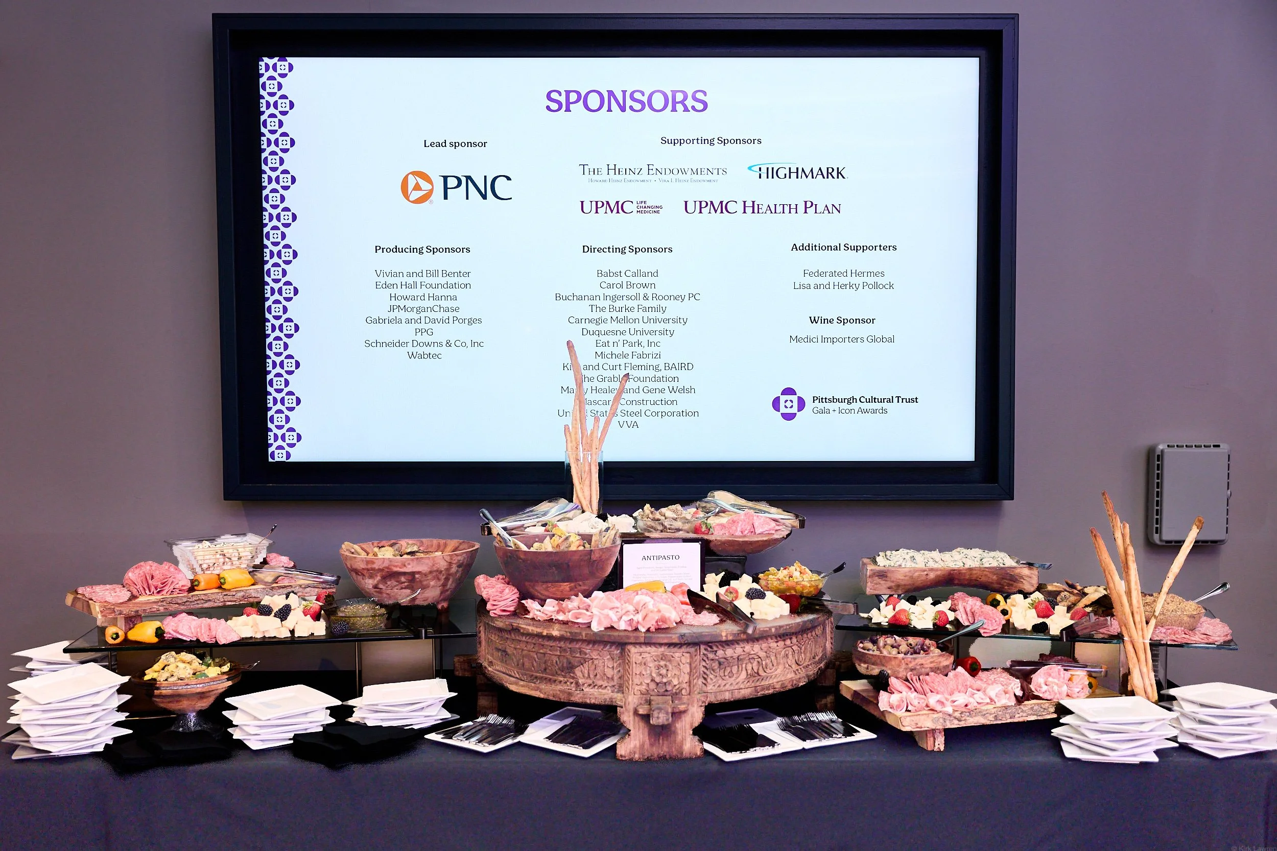 Dessert buffet with various sweet dishes and candies on black tables, with a digital board displaying sponsor information in the background.