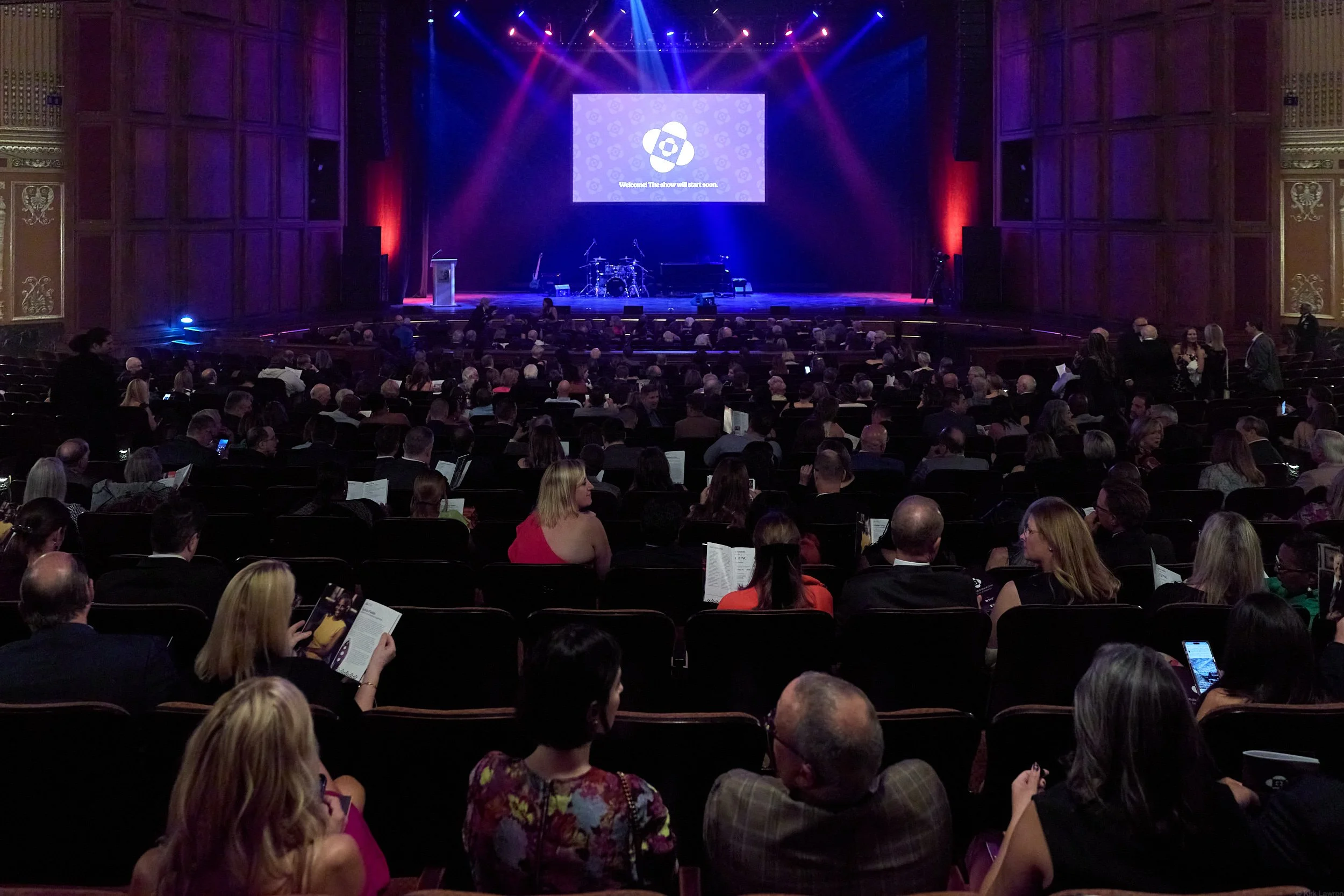 An auditorium filled with people waiting for a presentation, with a stage, musical instruments, and a large screen displaying a logo and the message 'Welcome! The show will start soon.'
