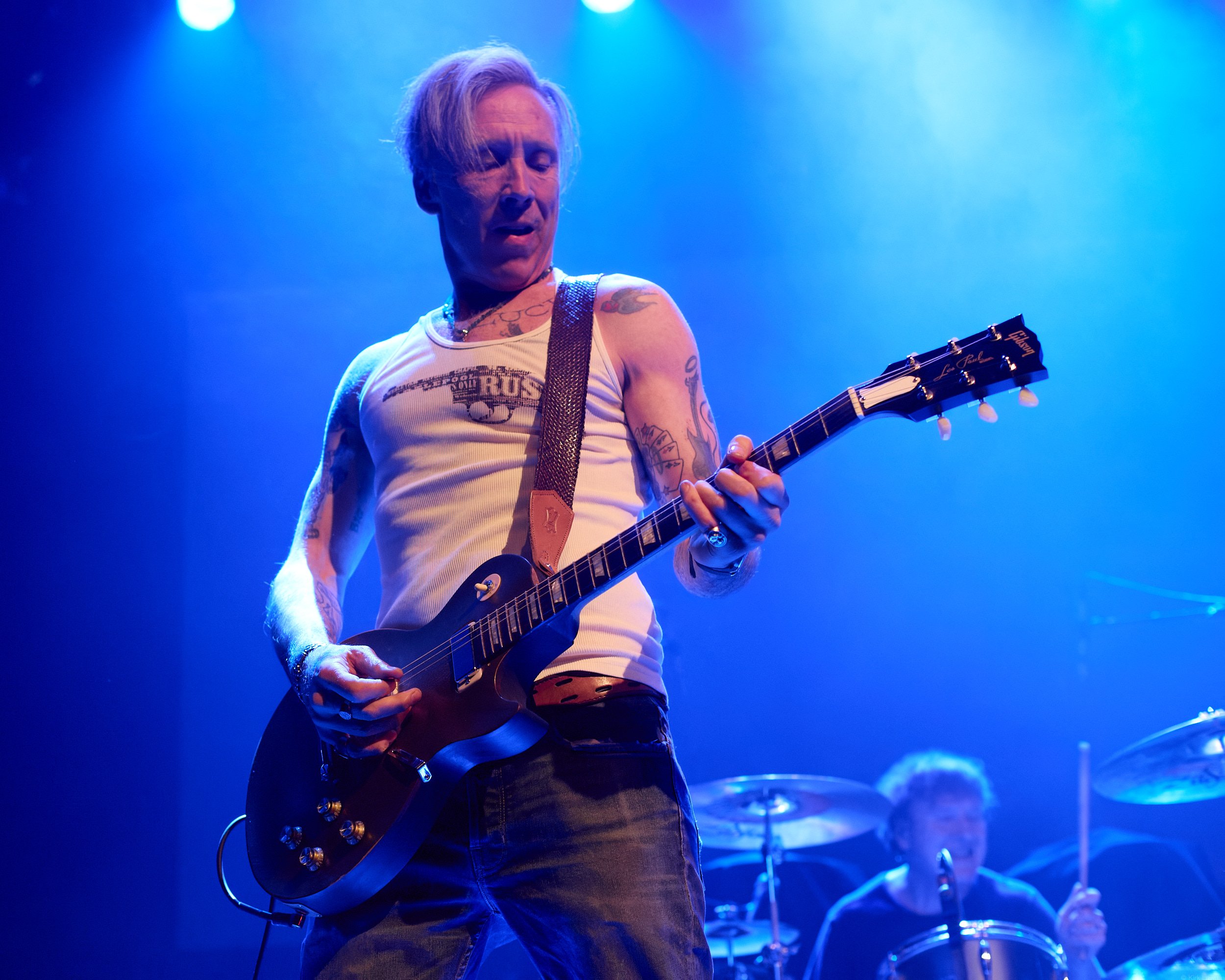 A male guitarist performing on stage, lit with blue and purple stage lights, wearing a sleeveless shirt, tattoos visible on his arms, and playing a Gibson Les Paul electric guitar.