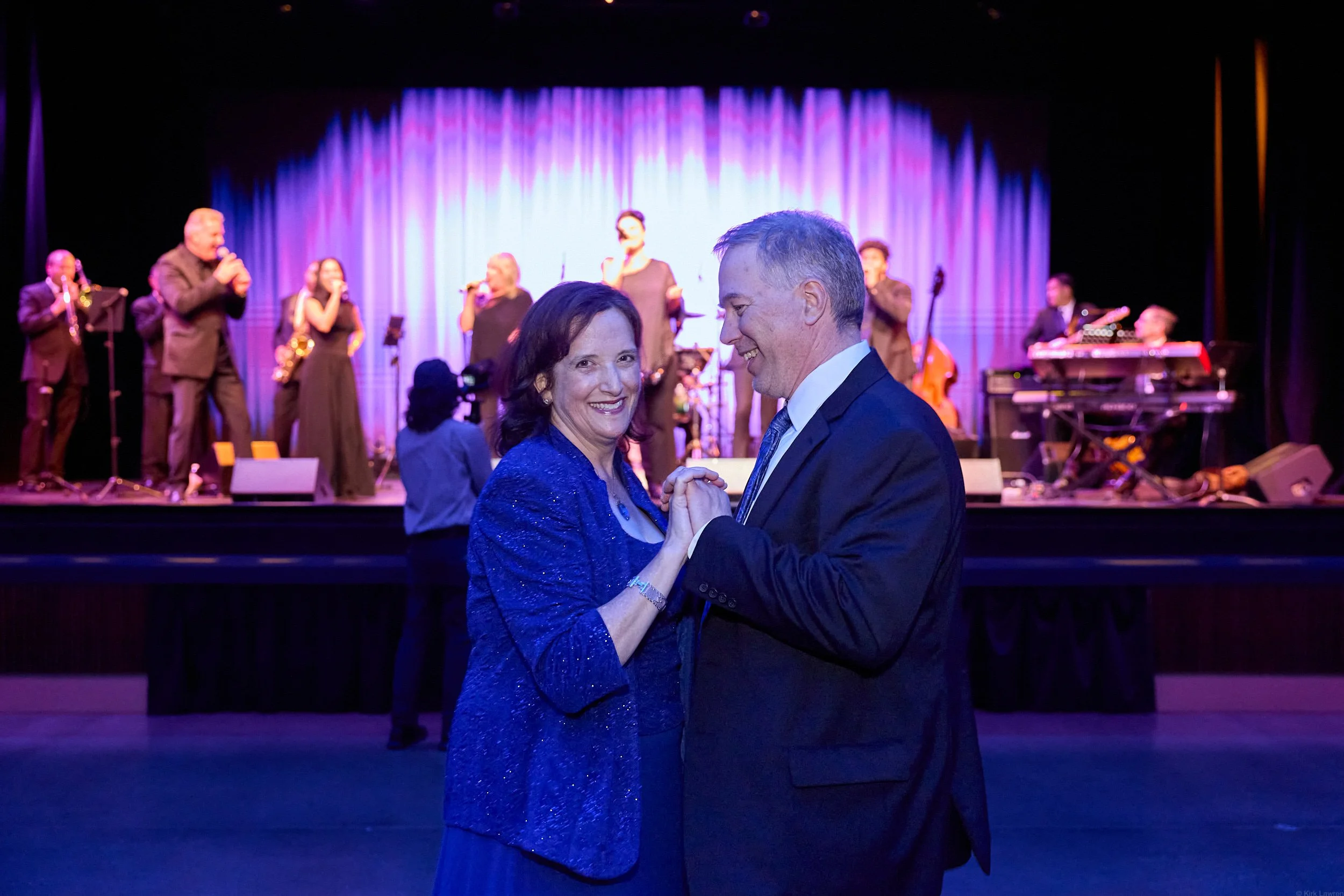 A couple dancing in the foreground at a formal event with a live band performing on stage in the background.