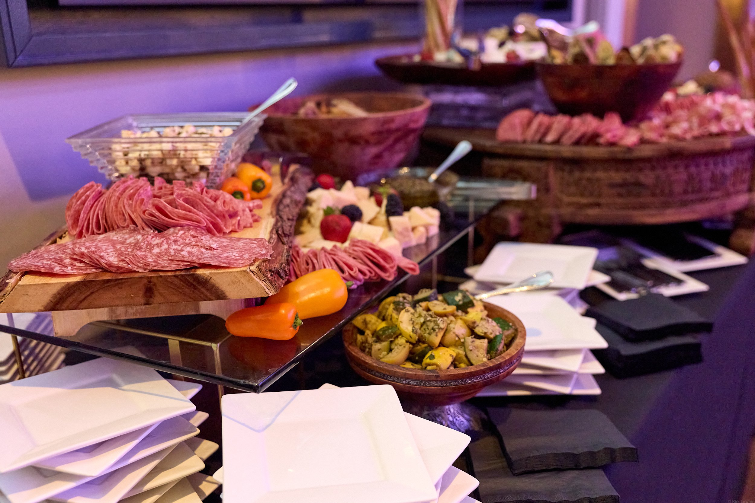 Buffet table with assorted cold cuts, cheeses, vegetables, and peppers on wooden and glass trays and bowls.