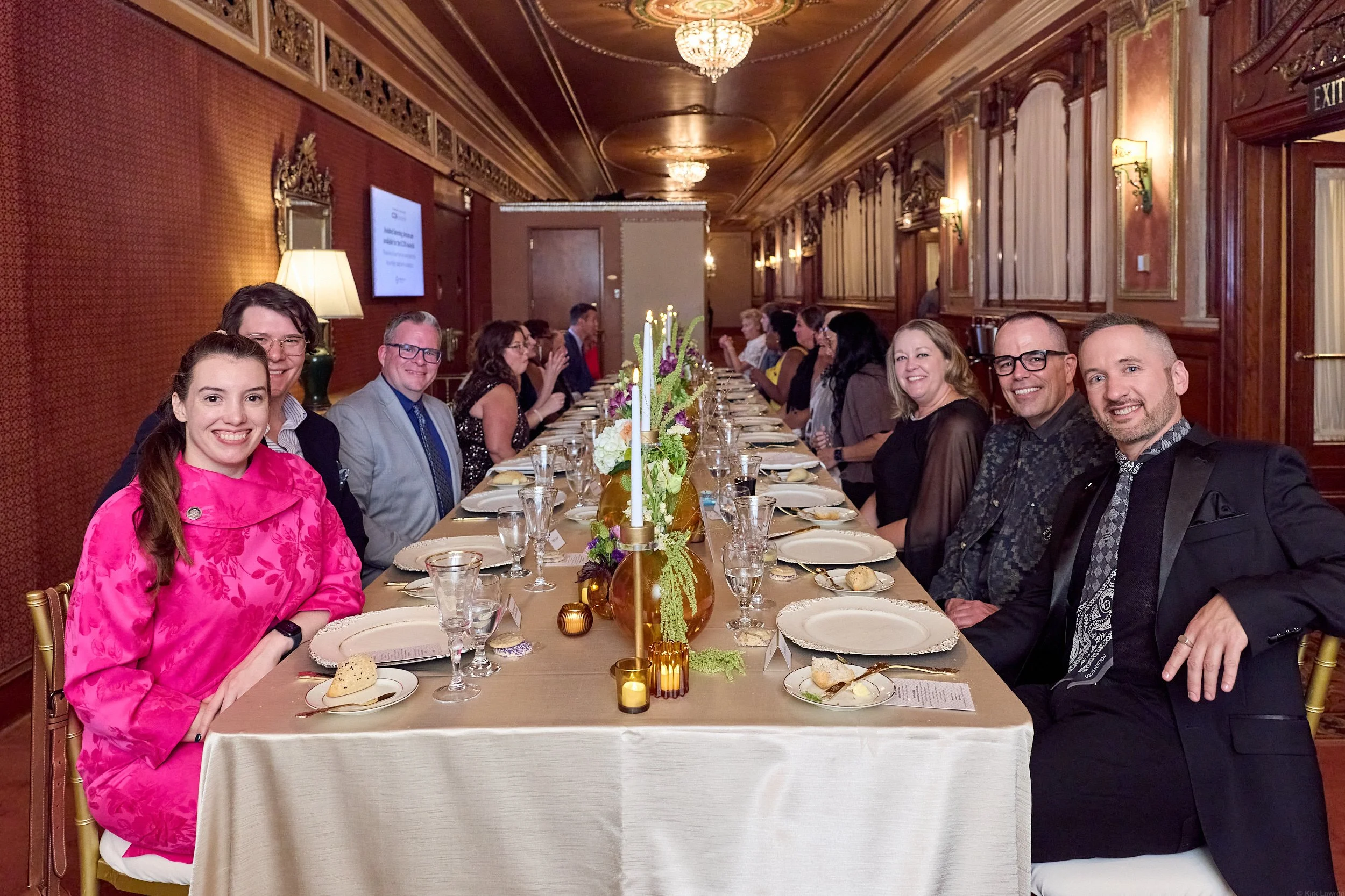 A group of people sitting at a long formal dinner table in a decorated room with chandeliers and ornate wood paneling. The table is set with plates, glasses, and a floral centerpiece with candles.