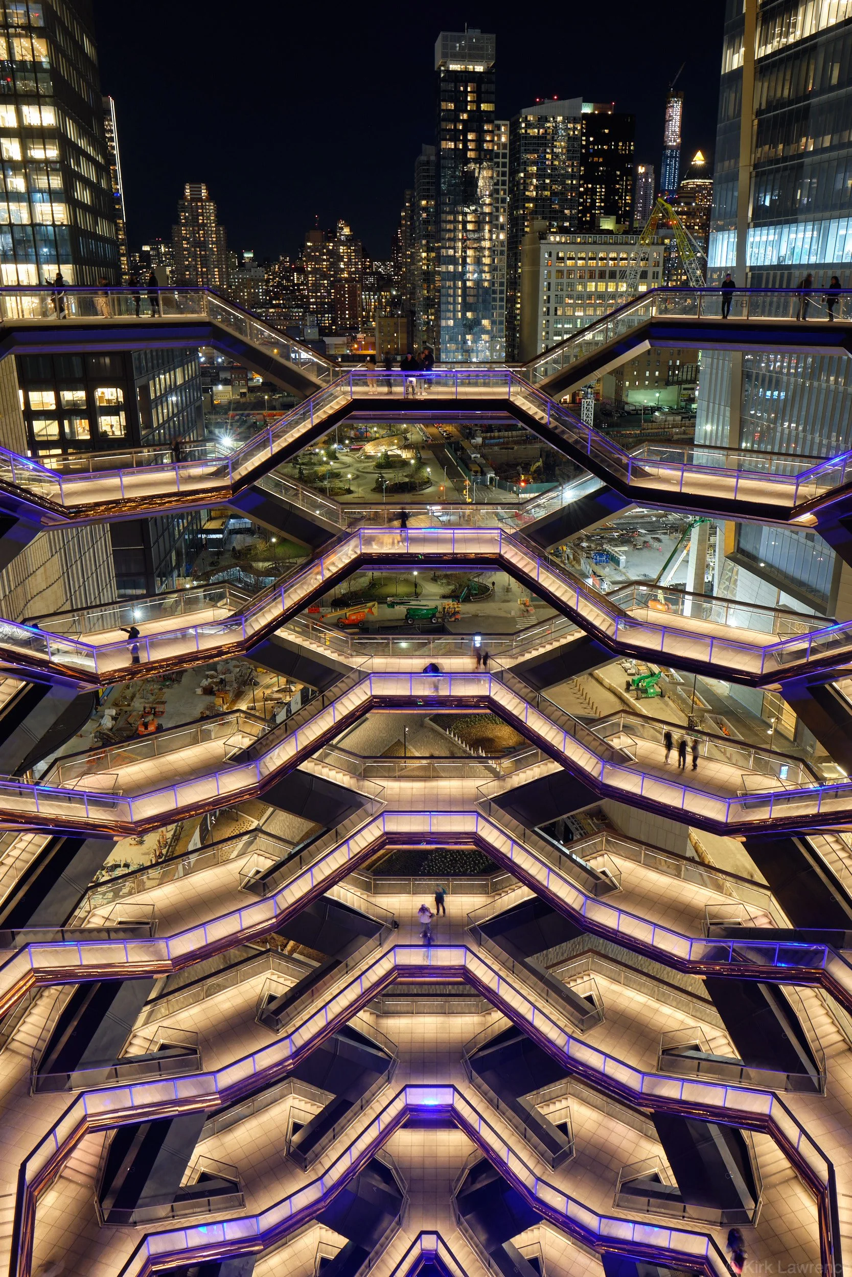 Nighttime view of a New York City cityscape from a high vantage point showing illuminated skyscrapers and modern architectural walkways with people walking, accented with blue lighting.