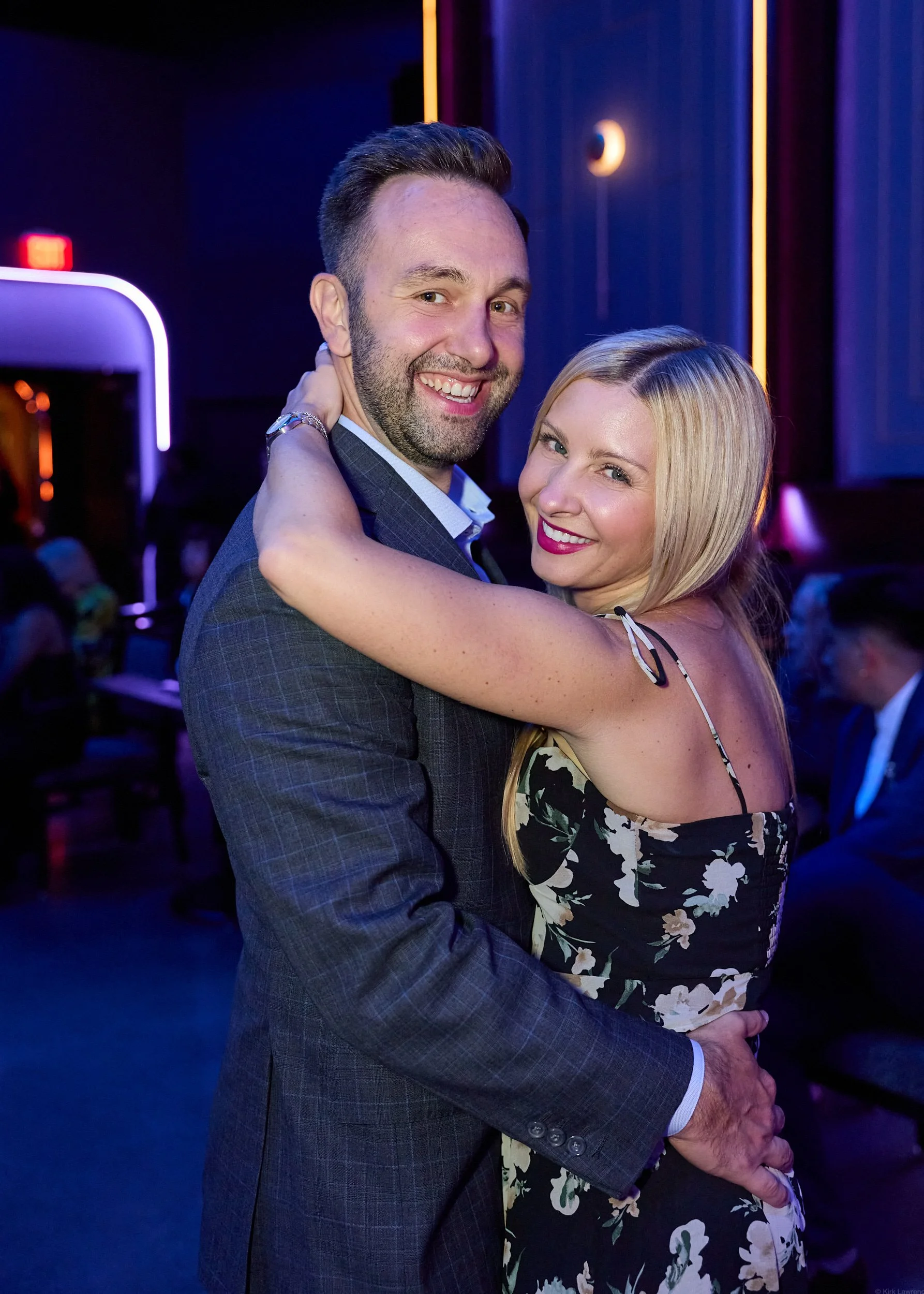 A man and woman dancing closely at an event, smiling, with a dark background and colorful lighting.