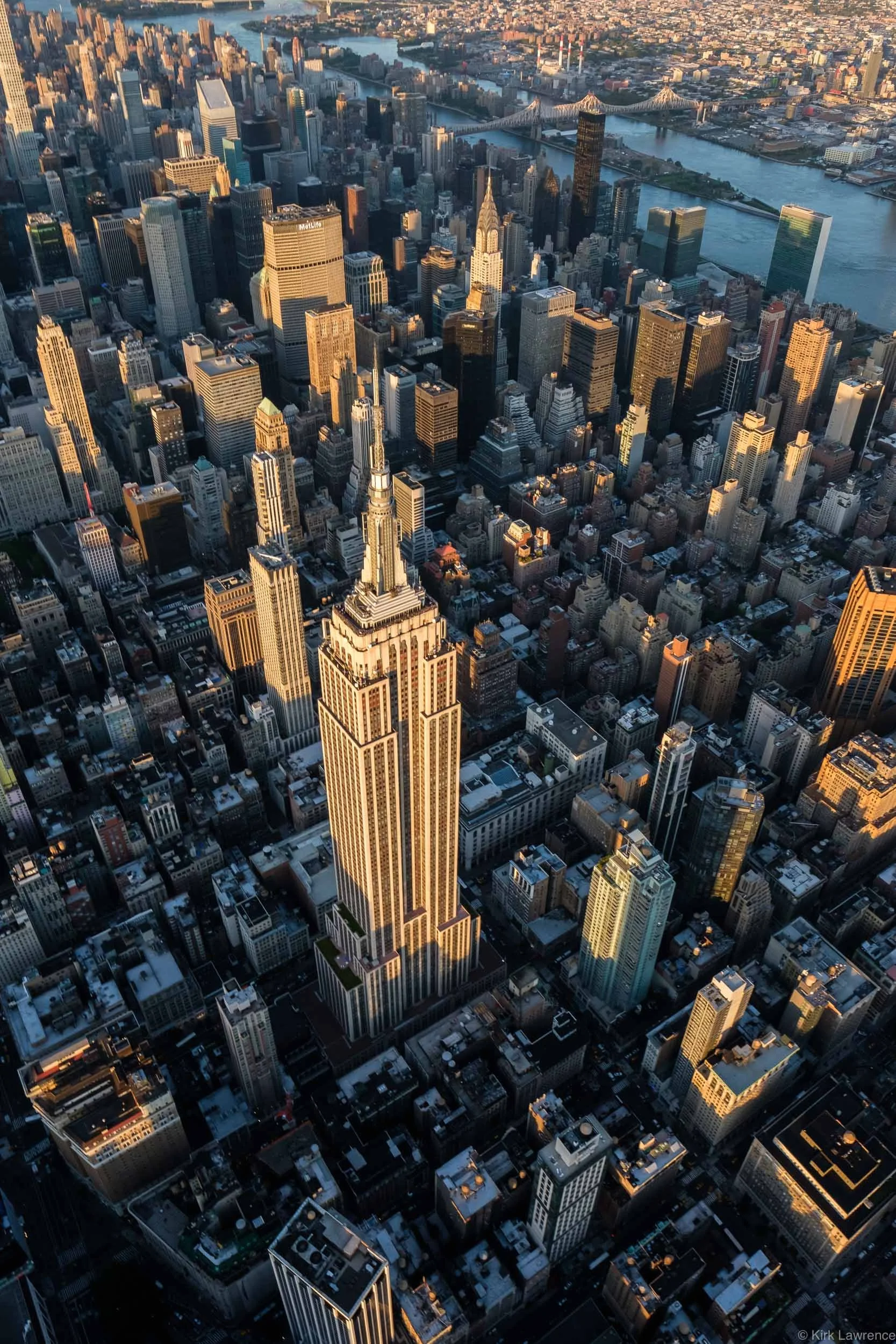 An aerial view of Manhattan in New York City during sunset, showcasing tall skyscrapers, the Empire State Building in the foreground, and the East River with bridges in the background.
