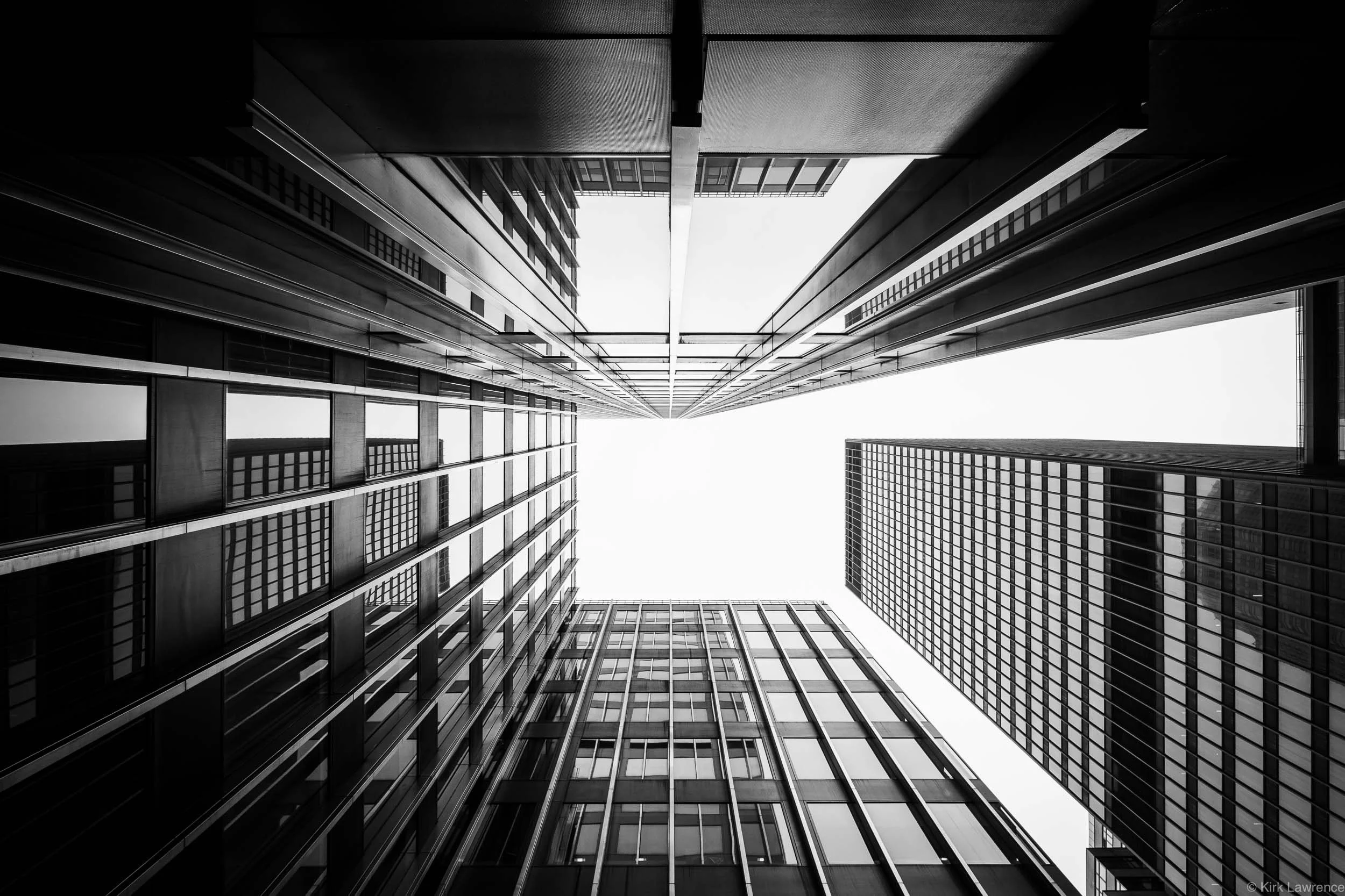 Black and white photograph of tall modern skyscrapers looking up from the ground to the sky, with reflections on glass windows.